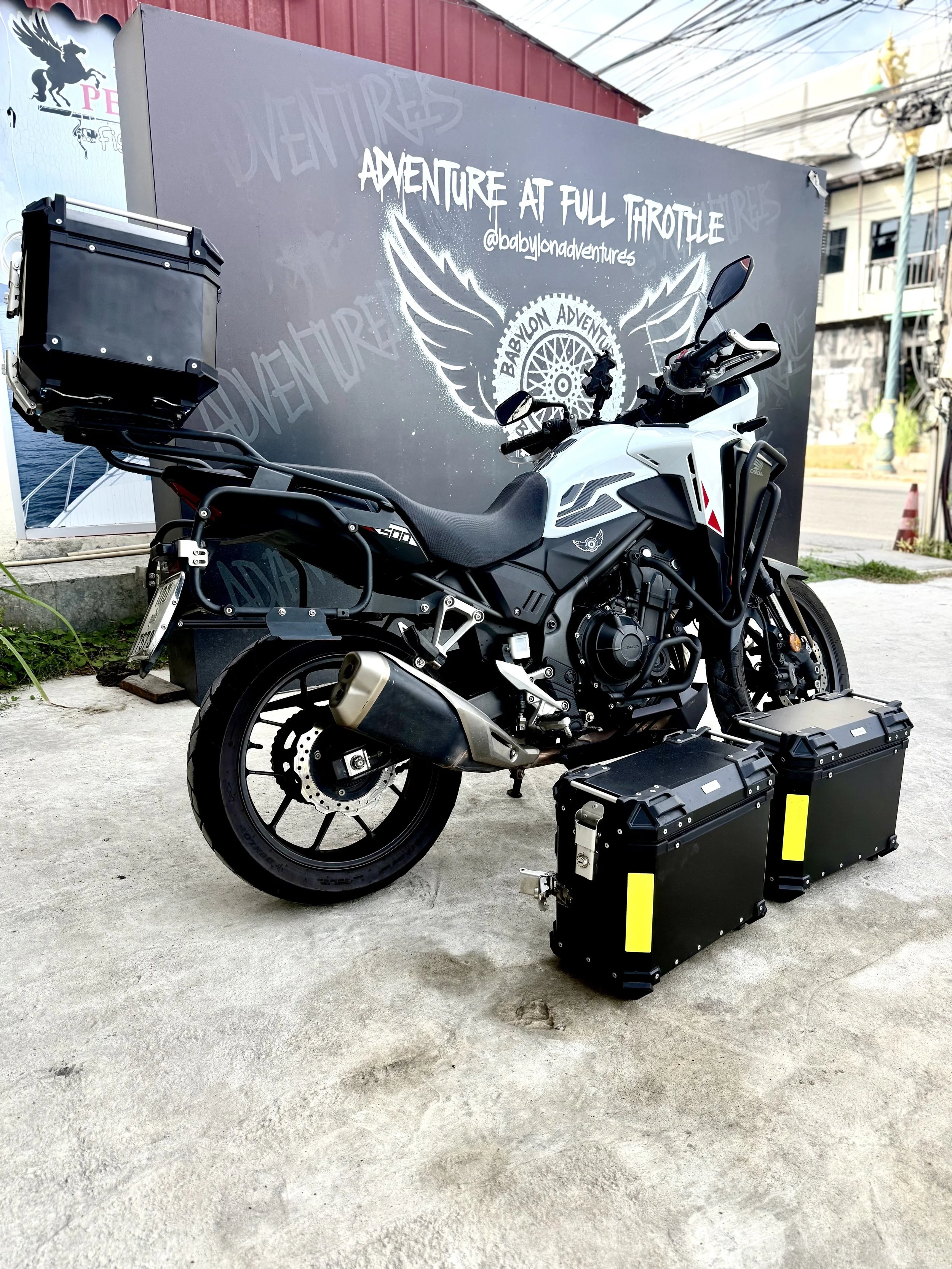 A white and black adventure motorcycle equipped with storage boxes on a concrete surface, with a blackboard behind it displaying the text 'Adventure at Full Throttle' and a logo for Babylon Adventures.