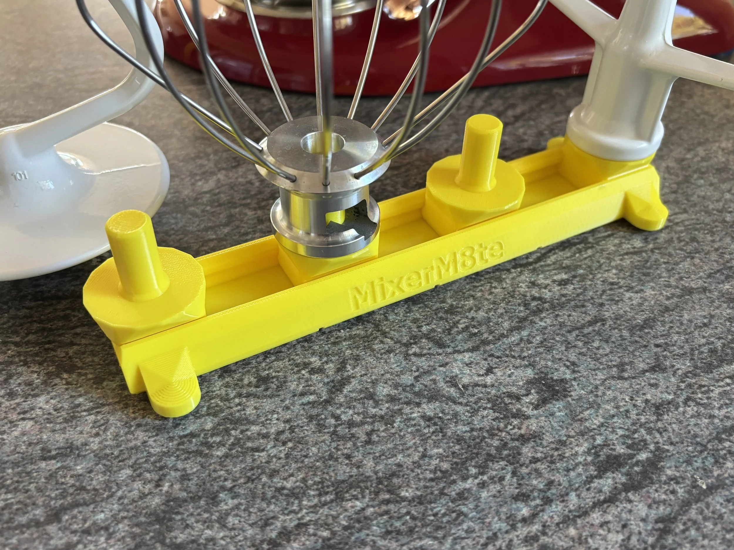 A yellow kitchen tool labeled 'MixerM8te holds a metal whisk attachment in place, with part of a white stand mixer visible in the background, all situated on a dark countertop.
