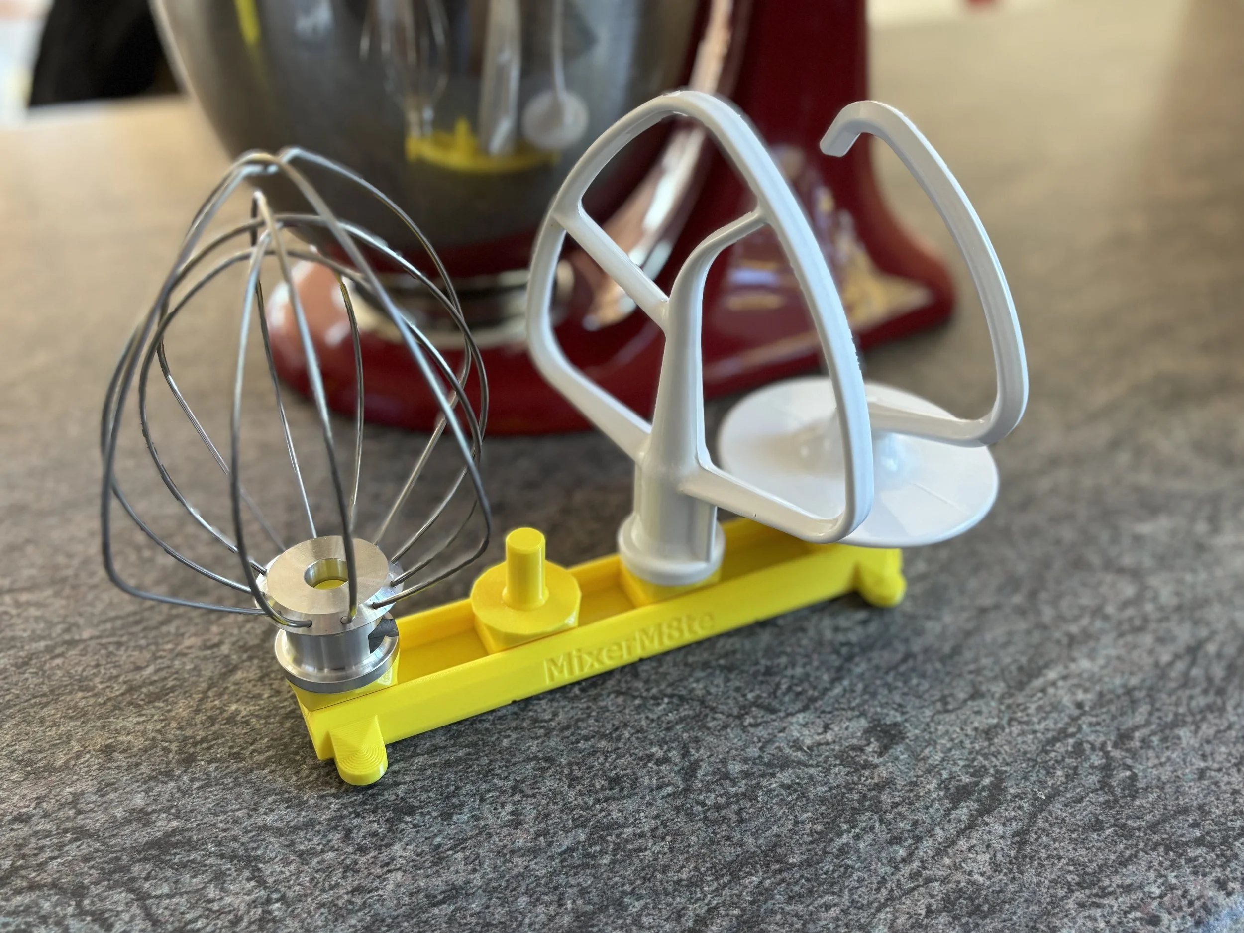 A yellow plastic MiniM8te accessories stand holding an electric mixer beater attachment  on a gray surface.