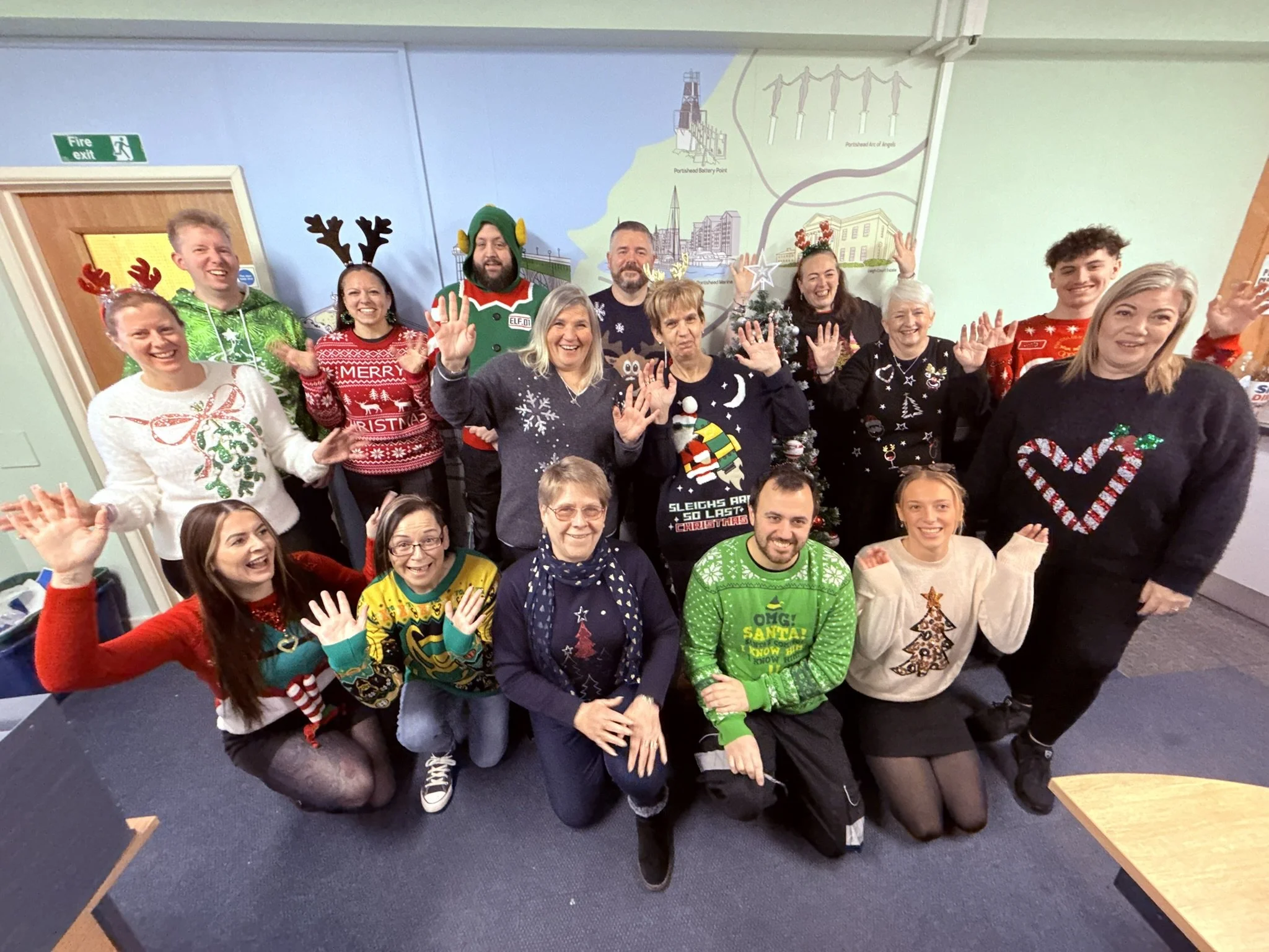 A group of people celebrating Christmas, wearing festive sweaters and holiday accessories, smiling and waving for a group photo indoors.
