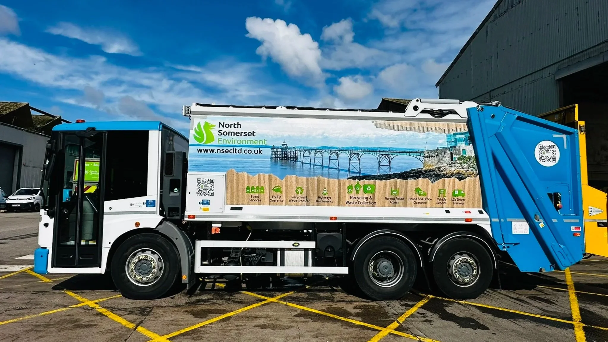 Side view of a recycling truck with "North Somerset Environment" branding in a parking lot, under a blue sky.