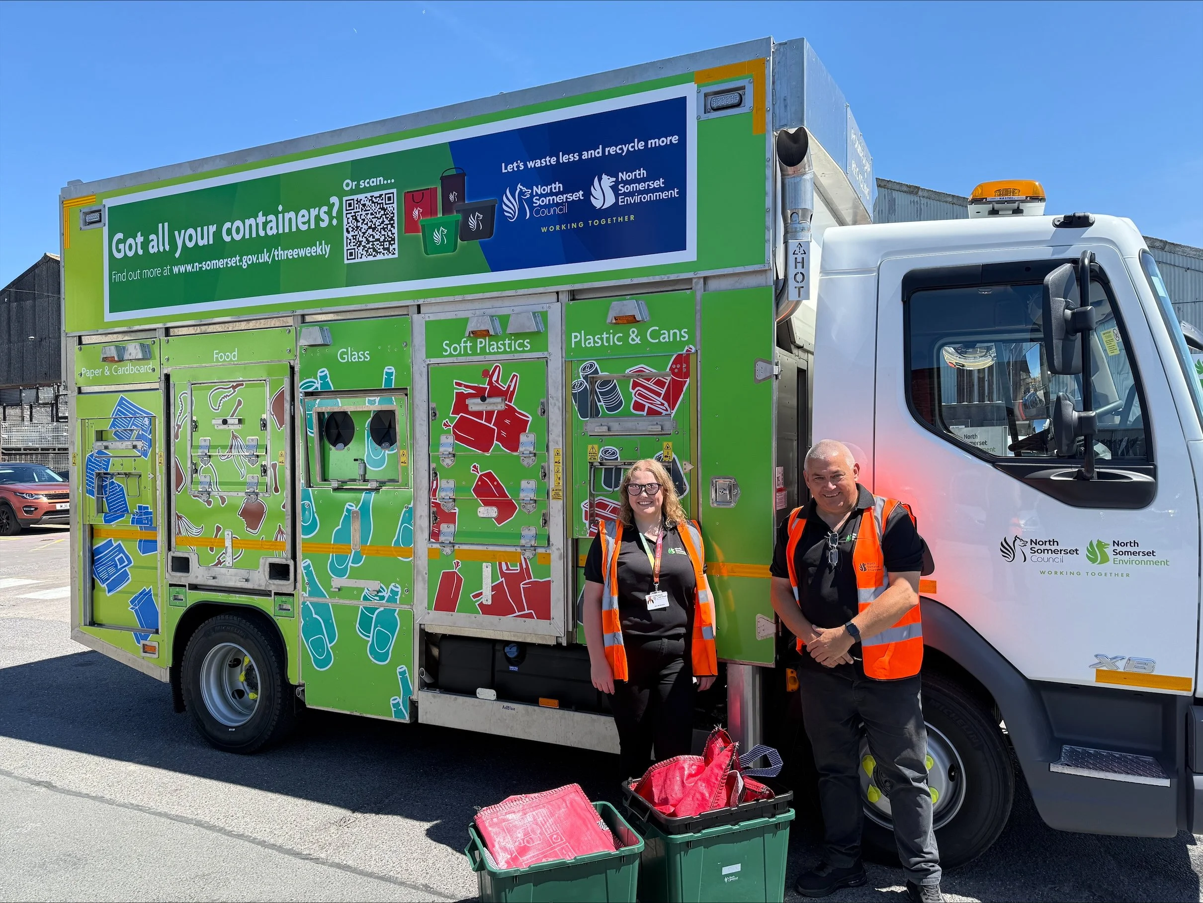Two recycling workers standing beside a green recycling truck with colorful graphics and the logos of North Somerset Council and North Somerset Environment. They are smiling and wearing orange safety vests, with recycling bins in front of them.