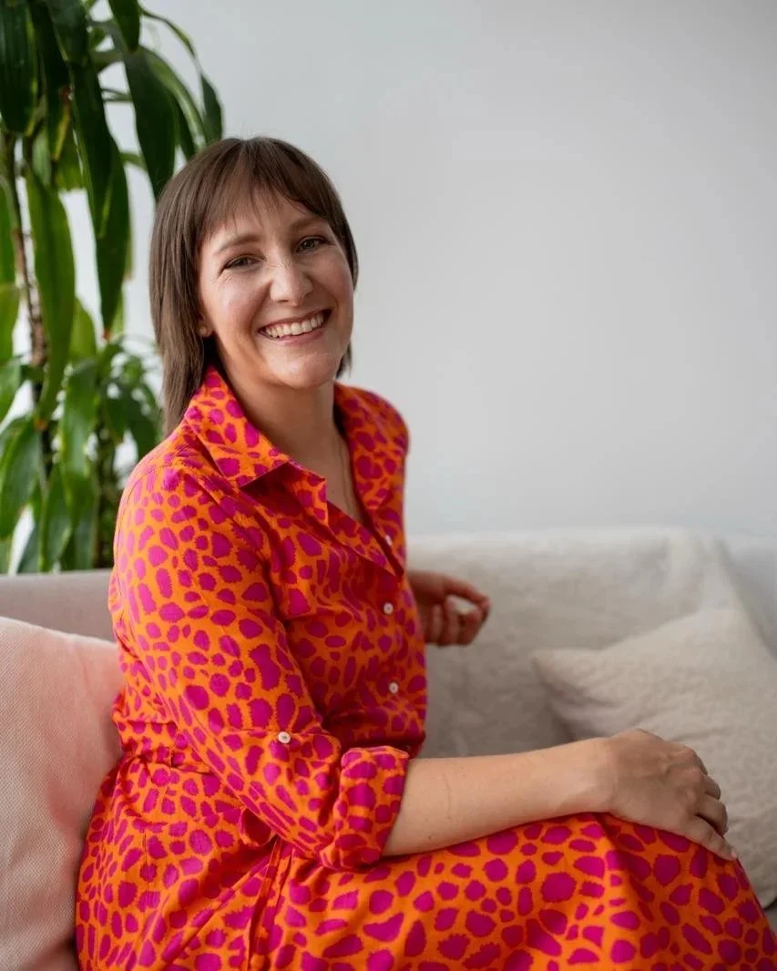 Porträt einer lachenden Frau mit kurzen dunklen Haaren, weißem Oberteil und pinker Jacke vor hellem blauen Hintergrund.