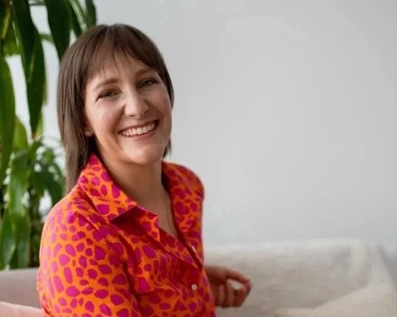 Lächelnde Frau mit kurzen dunklen Haaren, white T-Shirt und pinker Jacke vor hellem blauen Hintergrund.