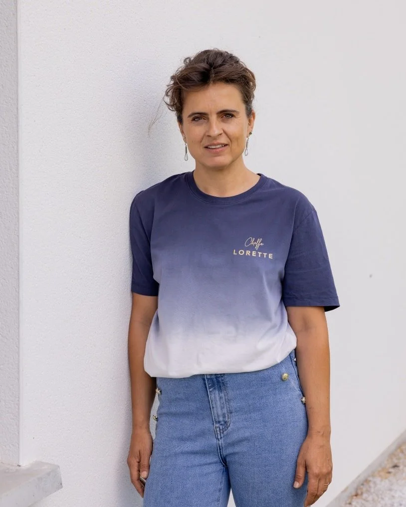 Une femme debout contre un mur blanc, portant une T-shirt bleu dégradé et un jean bleu, avec des boucles d'oreilles.