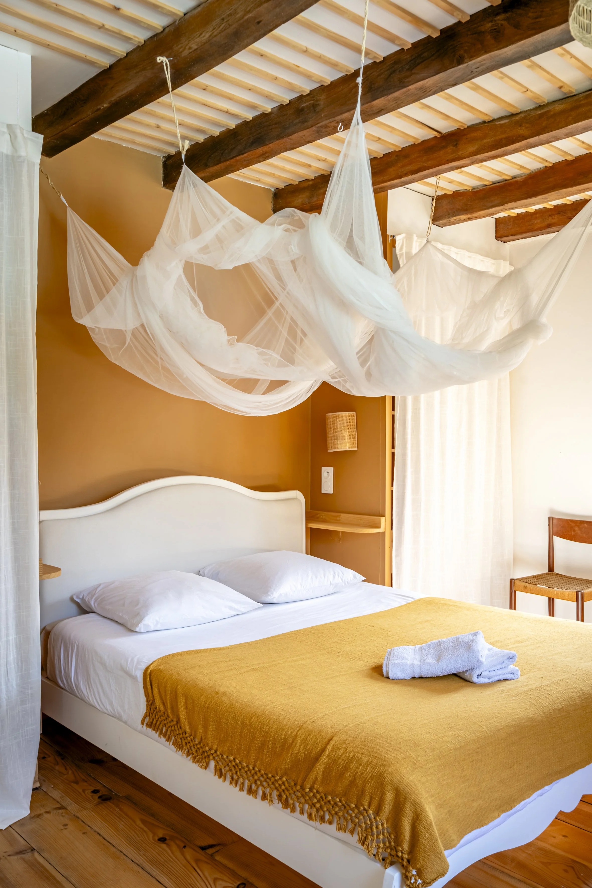 Chambre avec lit double, couverture jaune, serviettes blanches, lit en bois, poutres en bois au plafond, murs beige, rideaux blancs, support d'étagère en bois, chaise en bois, rideau blanc, décoration simple et chaleureuse.