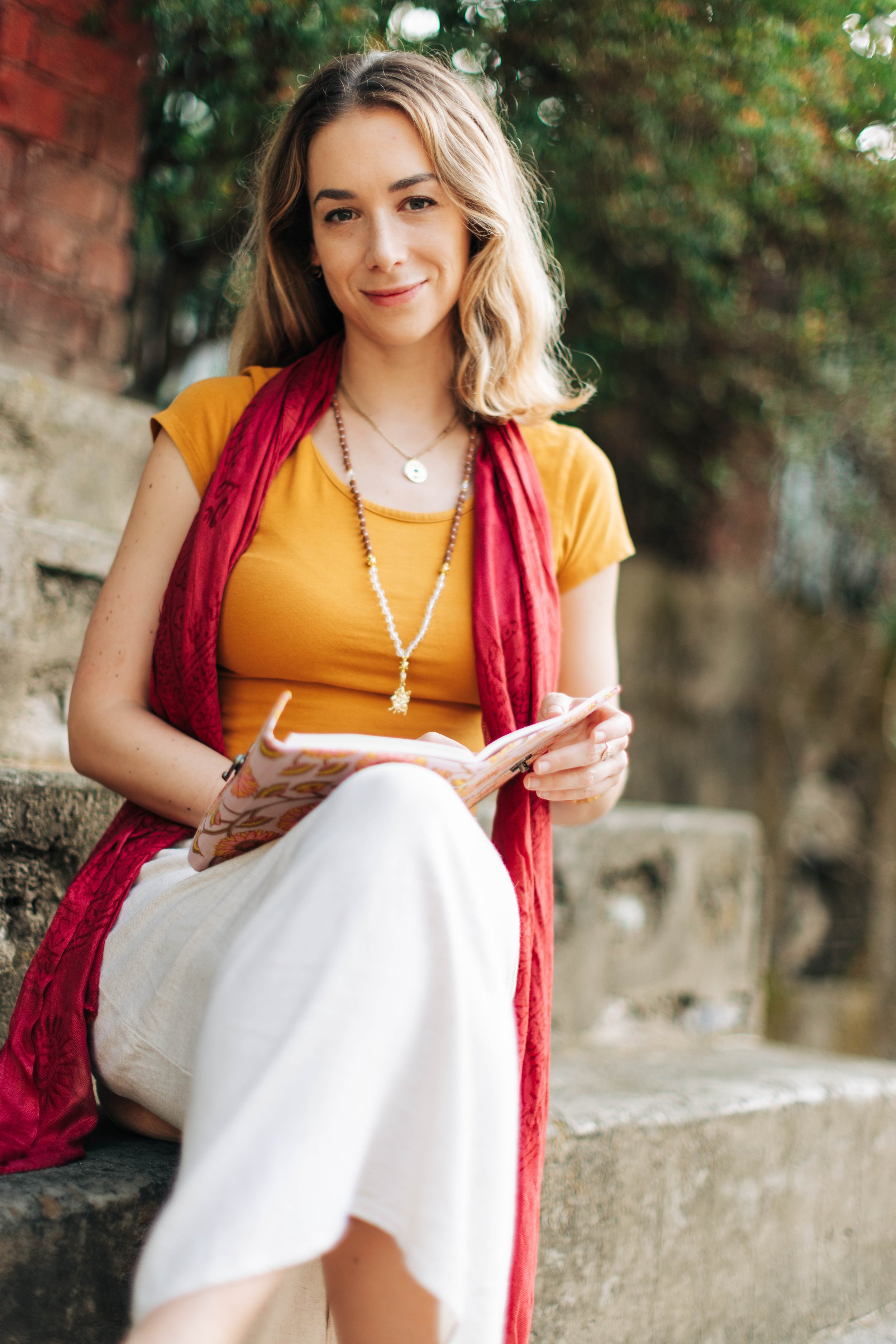 Une jeune femme assise sur un banc, souriant, portant un t-shirt jaune, un foulard rouge, un collier et un bracelet, tenant un carnet ou un journal.