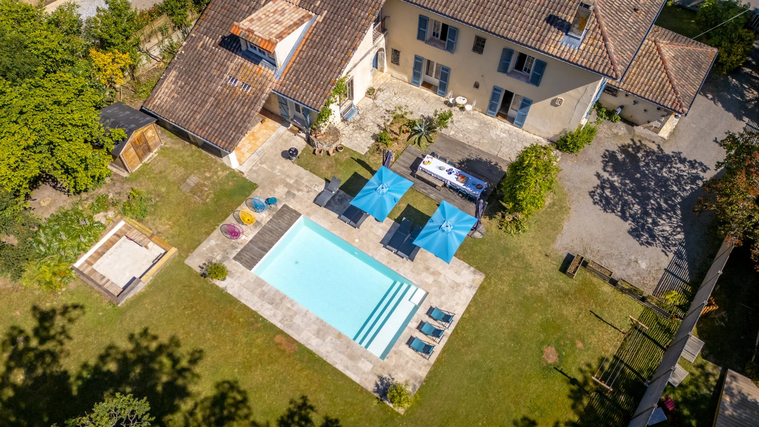 Vue aérienne d'une maison avec un patio, une piscine et une terrasse avec parasols bleus, entourée de jardins et de verdure.