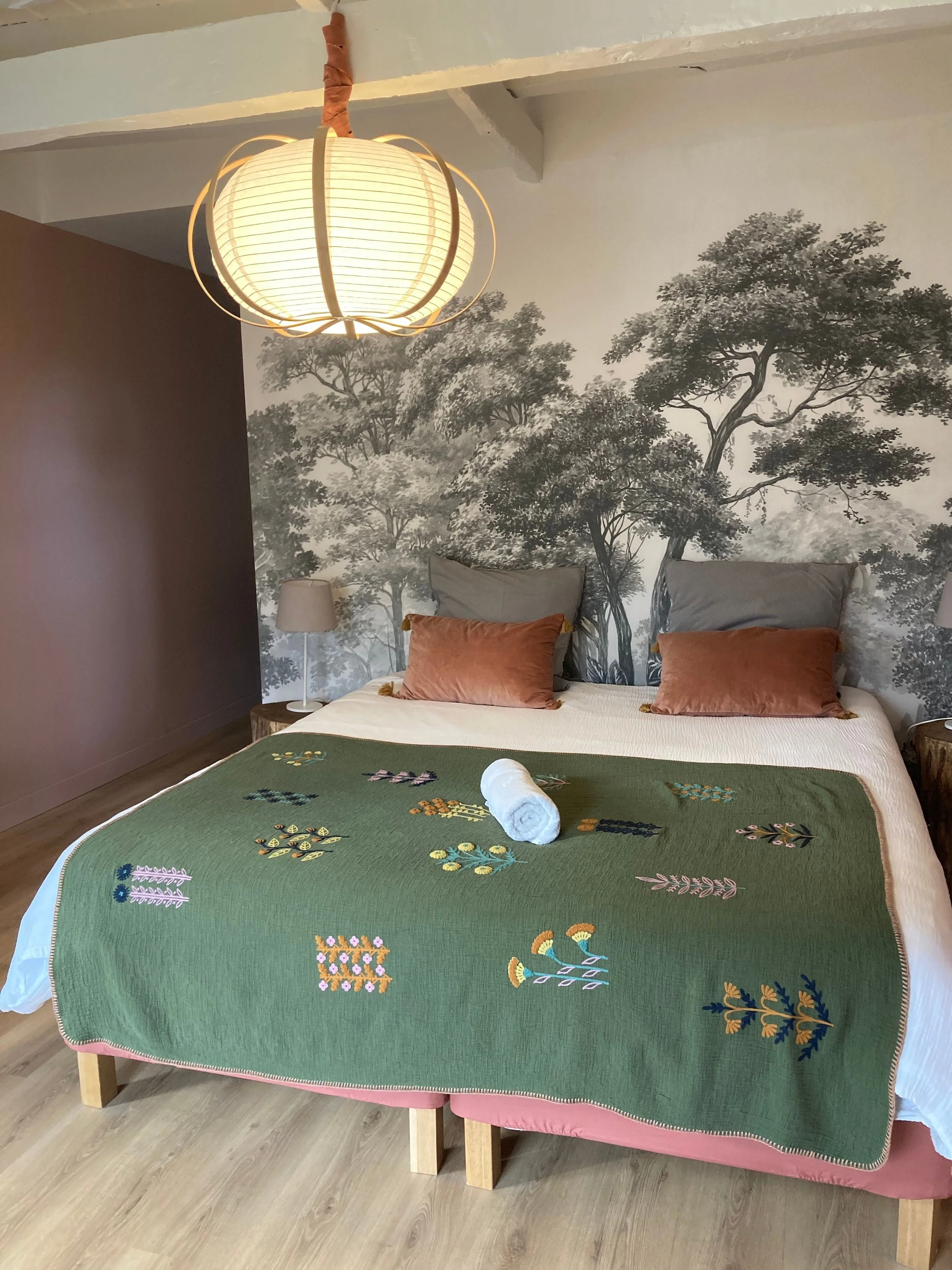 Un lit avec une tête de lit ornée d'un papier peint représentant des arbres en noir et blanc. Le lit est recouvert d'une couette verte avec des broderies florales, et il y a deux coussins gris et deux coussins en velours roux. Sur le lit, un rouleau de serviette en papier blanc est placé.
