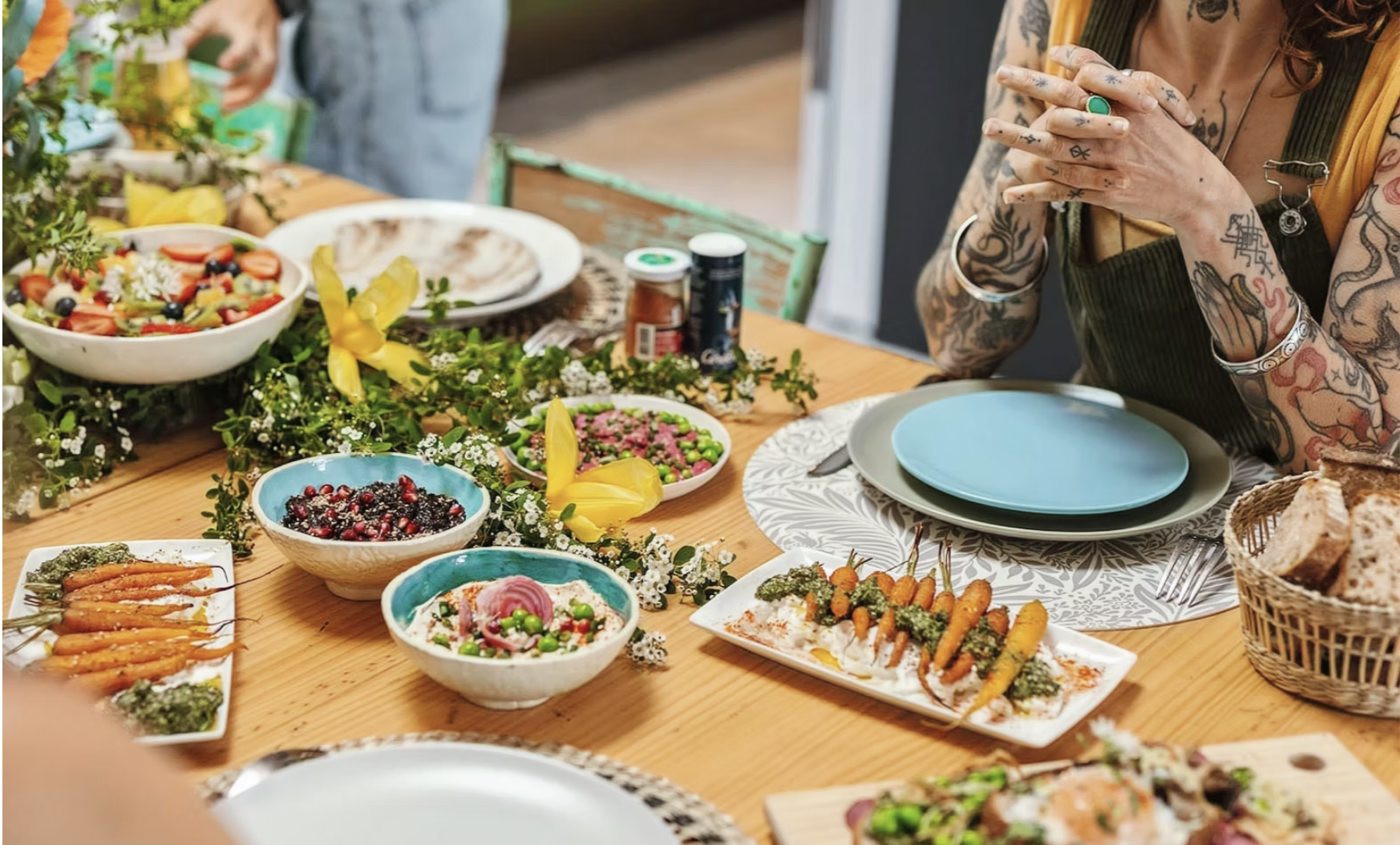 Vue d'une table de repas avec plusieurs plats colorés, des salades, légumes grillés, et fleurs décoratives, avec une personne tatouée assise à la table.