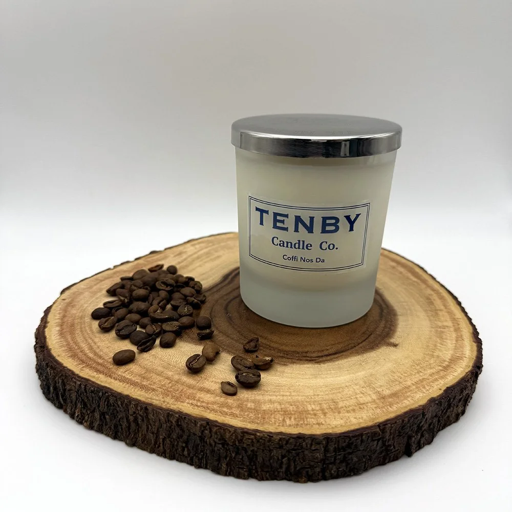 A white candle with a silver lid and a label that reads "TENBY Candle Co." placed on a round wooden board with coffee beans beside it.