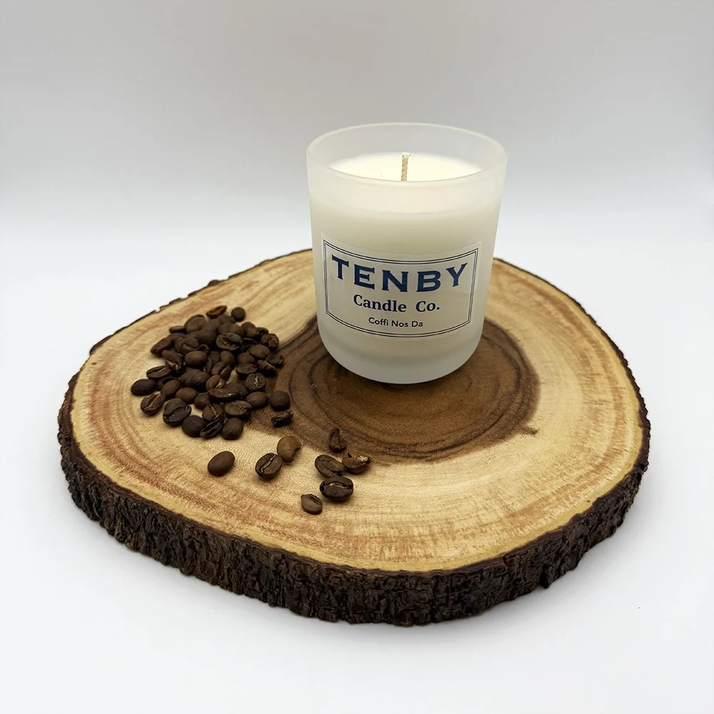 A white candle in a glass jar labeled "Tenby Candle Co." placed on a round wooden slab with coffee beans beside it.