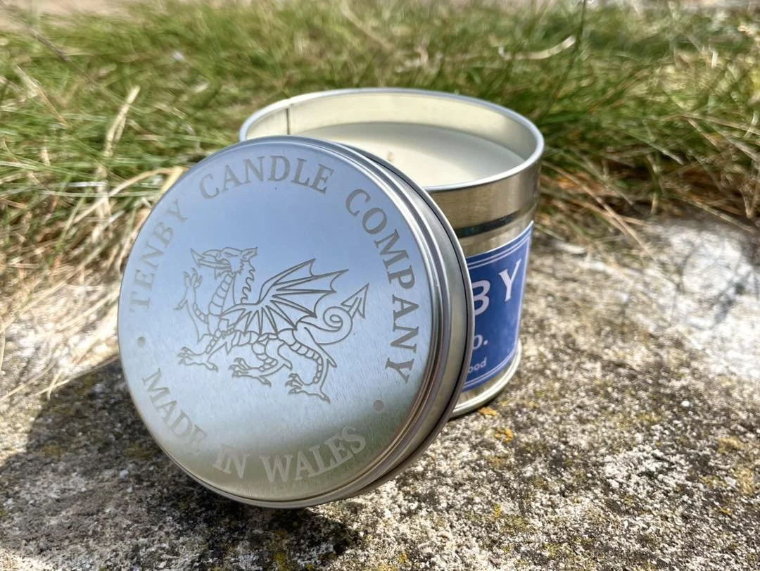 A tin of scented candle from Tyenb Candle Company placed on a concrete surface with green grass in the background.