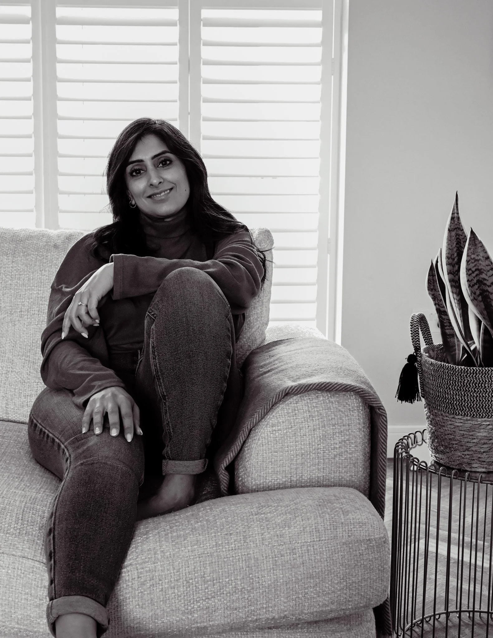 Black and White image of lady sat on sofa with one leg bent, smiling at the camera