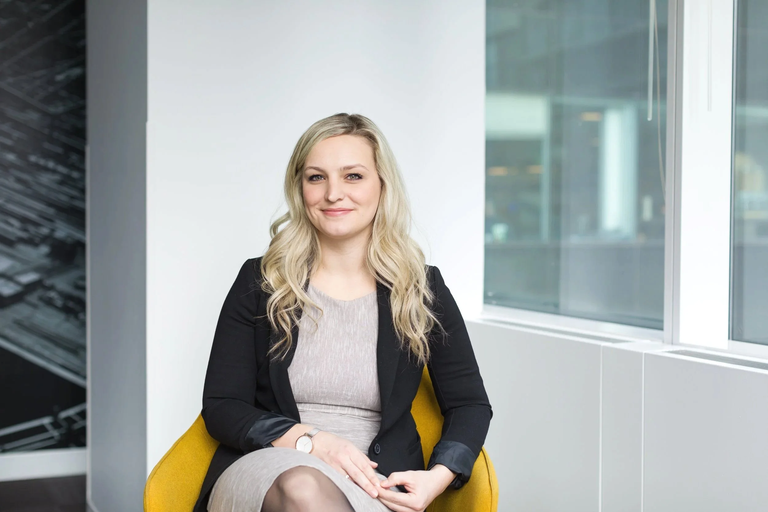A branding shots of a female employee with blonde hair and a blazer sitting in a yellow chair at her office 