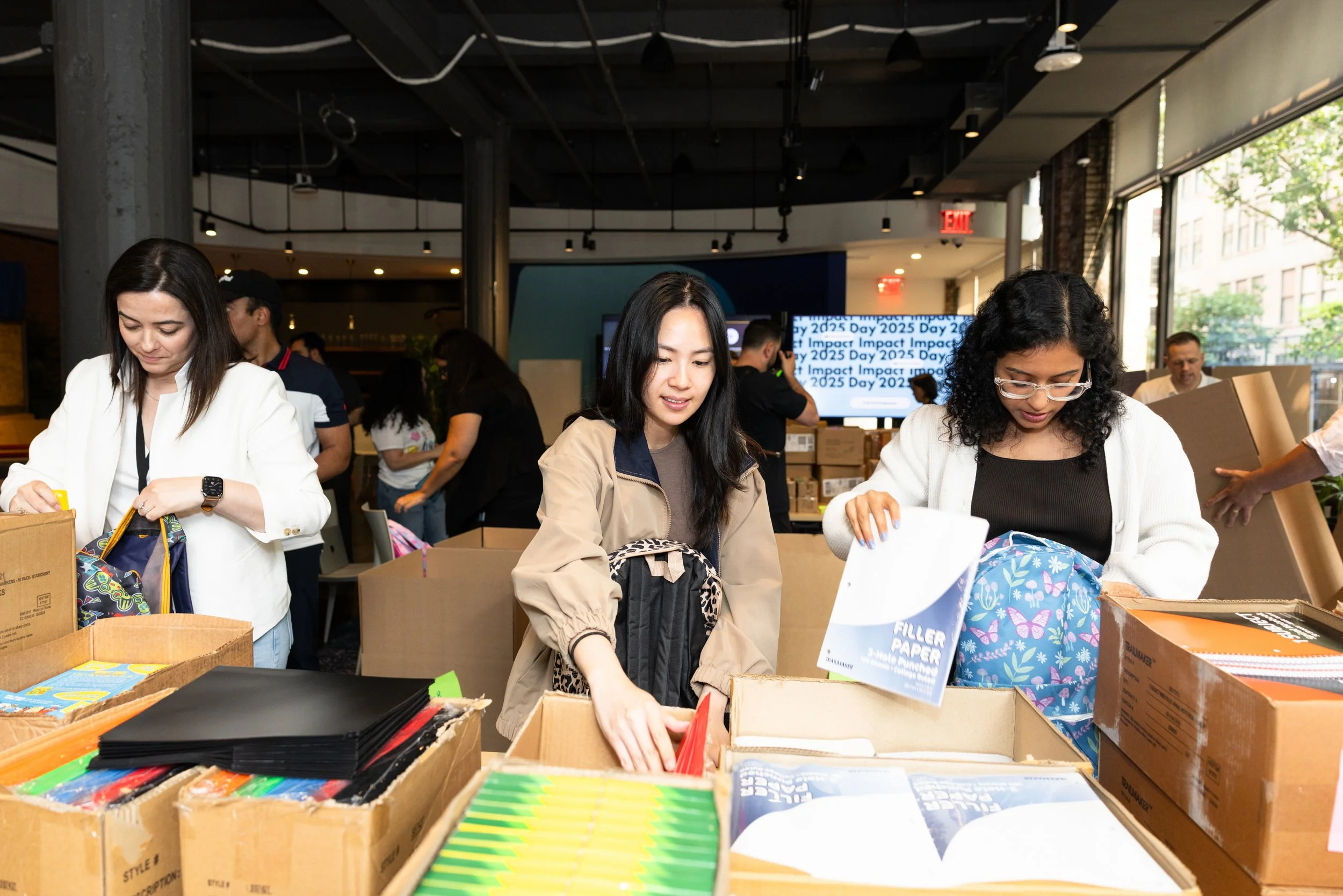 three women stuffing backpacks with school supplies sorted in boxes with other volunteers behind them