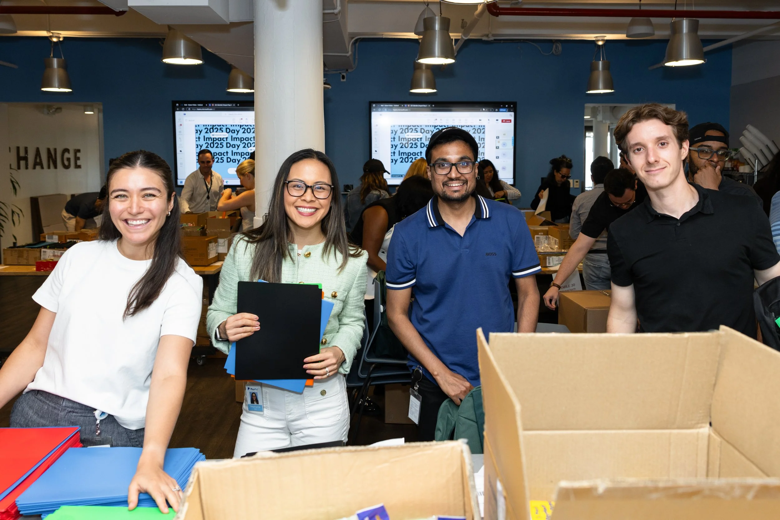 four volunteers smile at the camera in their offices as they stuff backpacks with school supplies