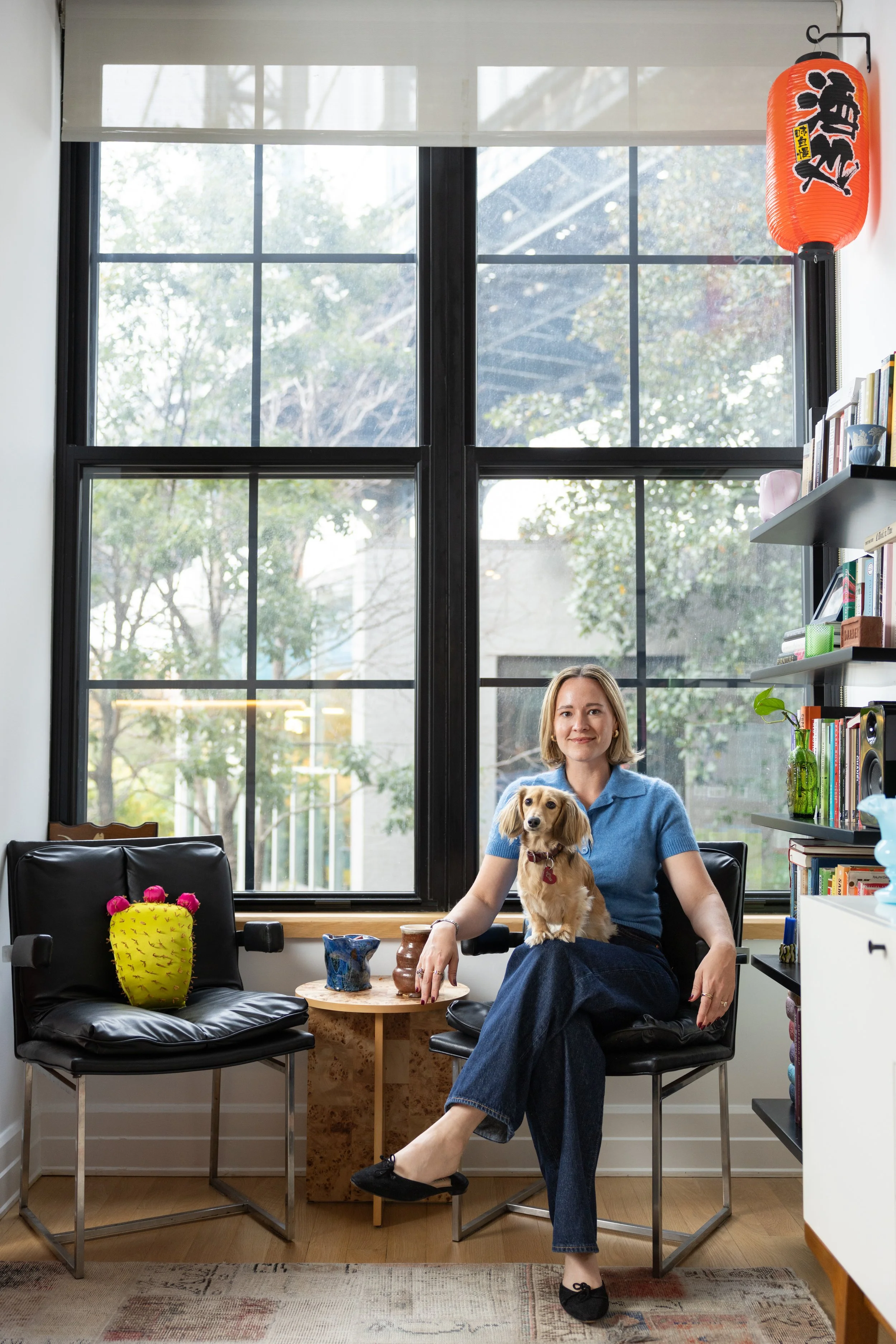 A woman sits with a dog on her lap in front of a window with a view of the Manhattan Bridge 