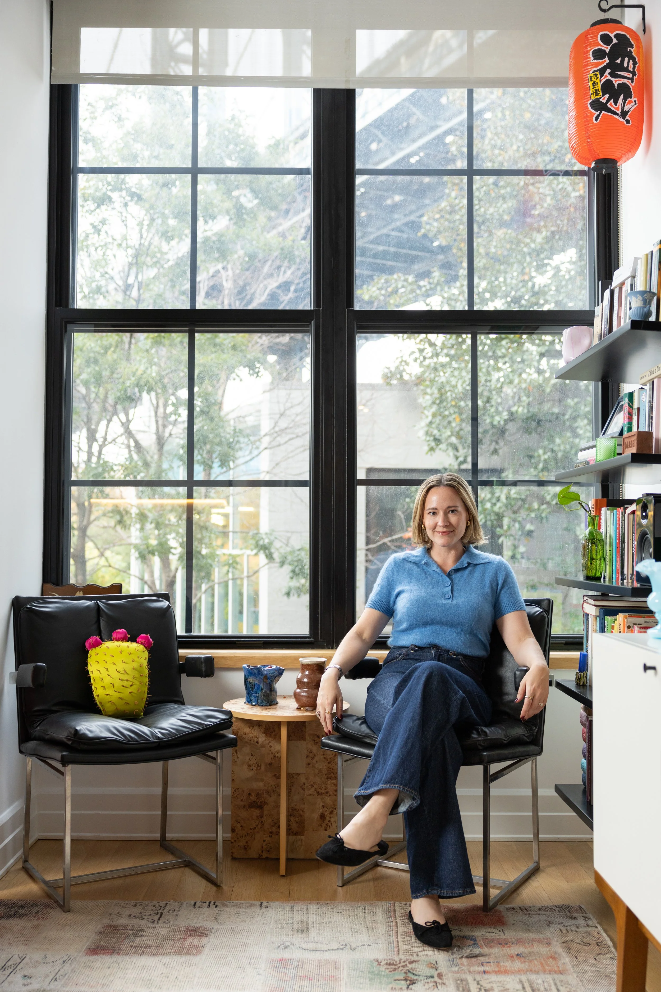 A woman with blond hair and a blue shirt and jeans sits in casually in a chair in her New York apartment