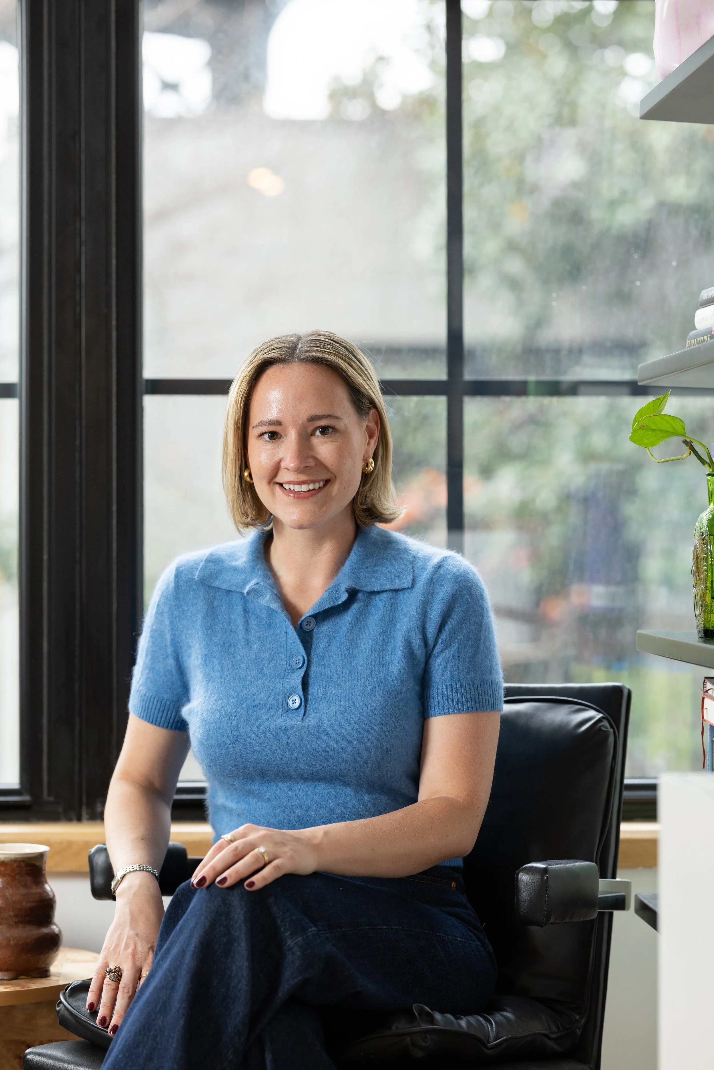 A woman with blond hair and a blue shirt sits in a black chair smiling at the camera in her New York apartment