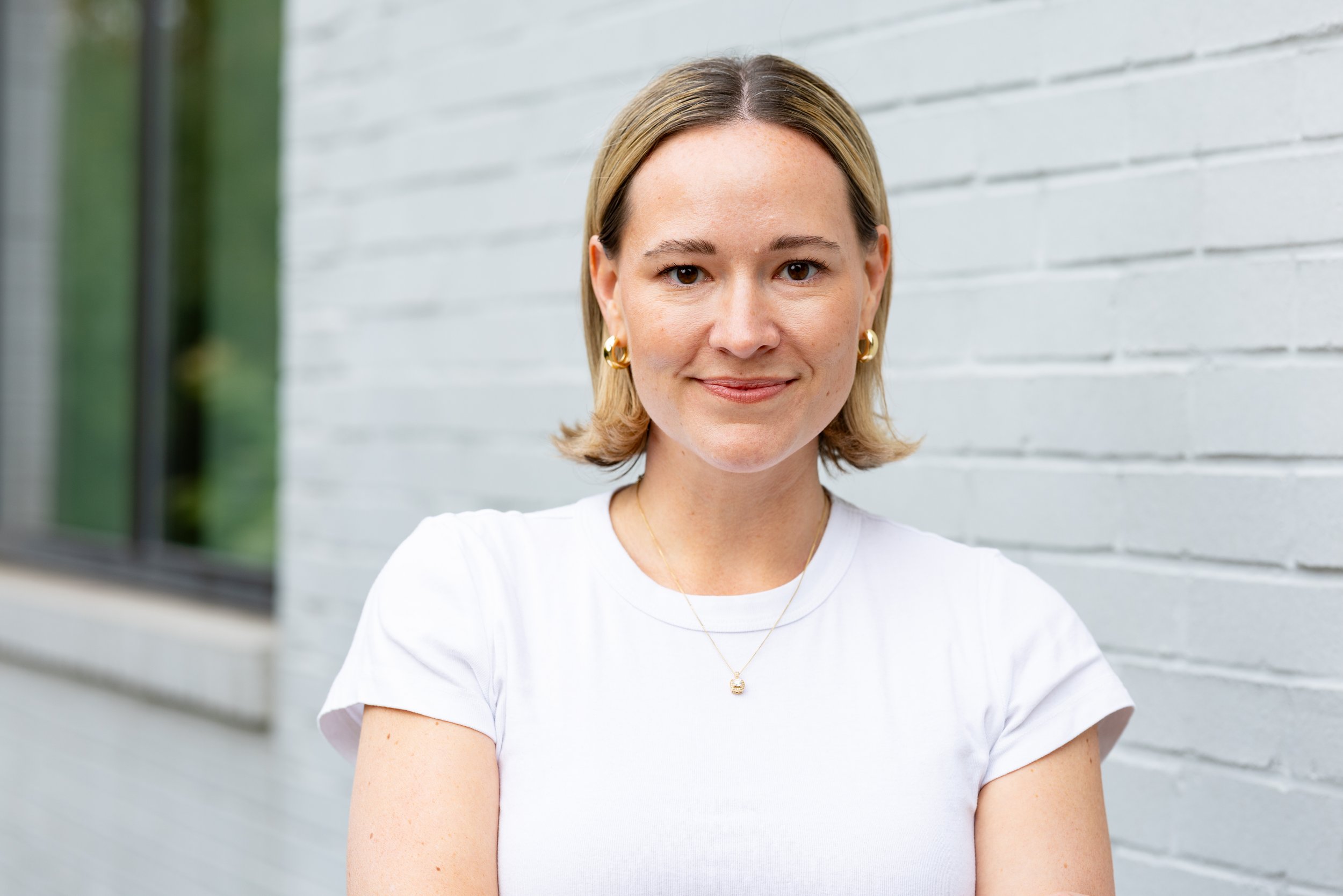 A woman with a blond bob and white t shirt stands in front of a white brick wall