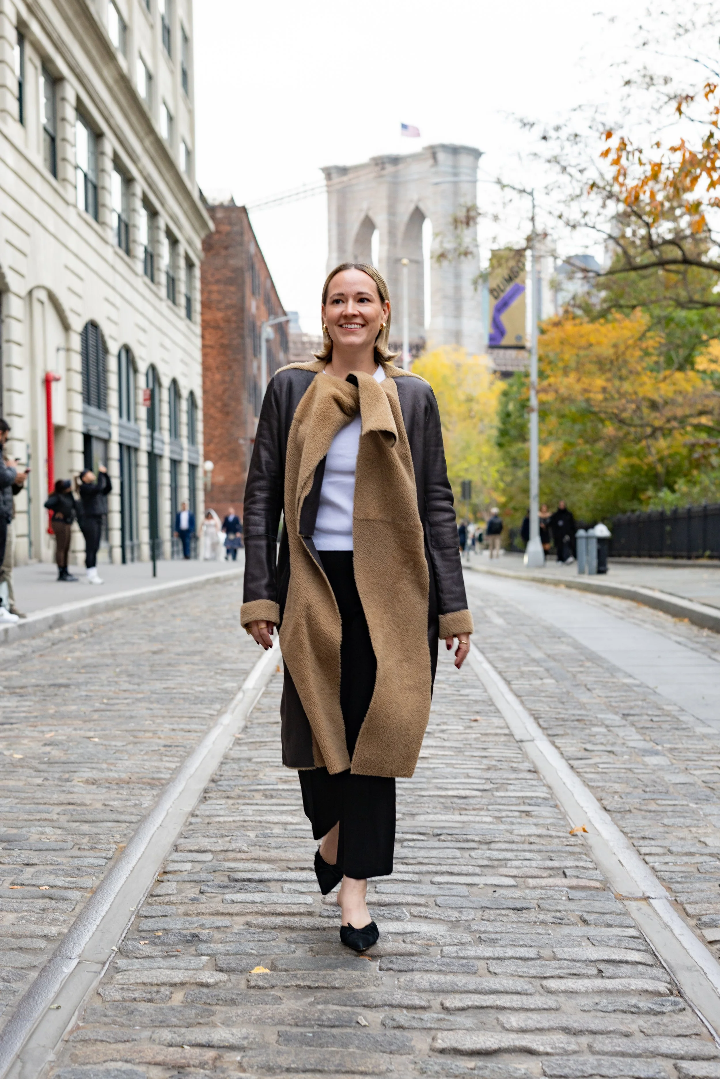 A woman walks confidently wearing a leather jacket on a cobblestone street with the Brooklyn Bridge in the backdrop