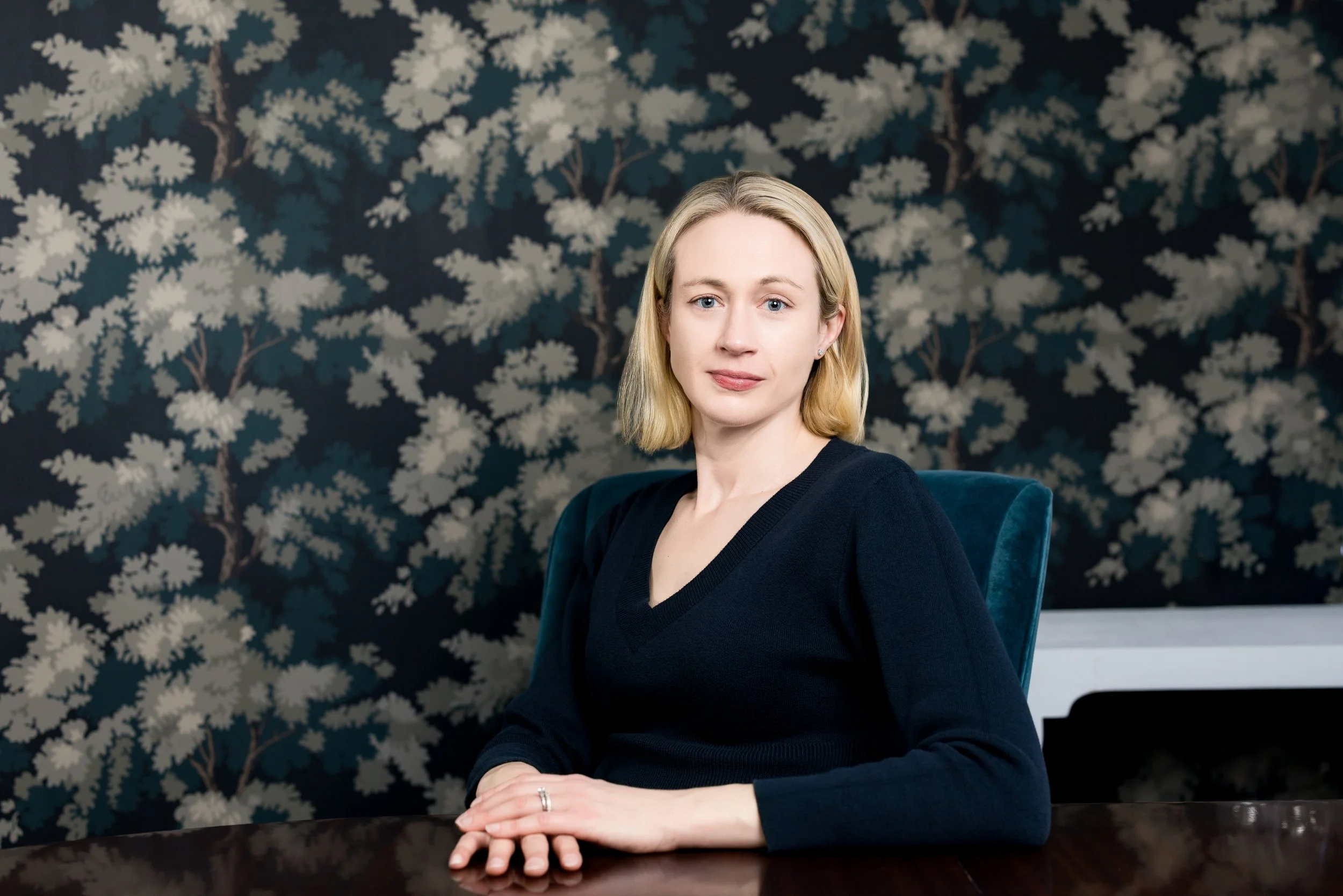 Portrait of a woman with a v neck sweater and blond hair looking at the camera leaned on a table with dark wallpaper behind her