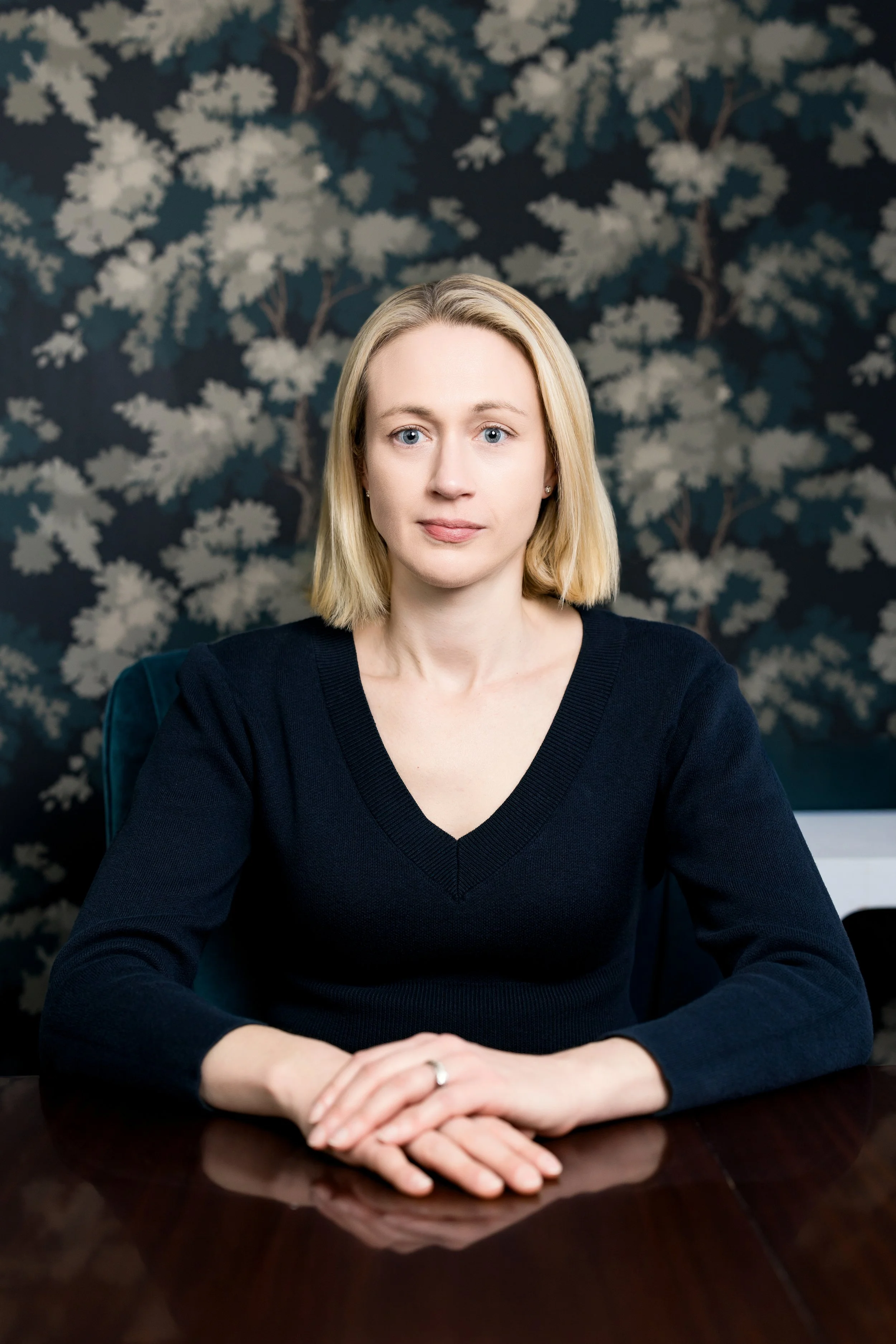 Blonde woman with blue eyes looking at the camera seated at a table with wallpaper behind her