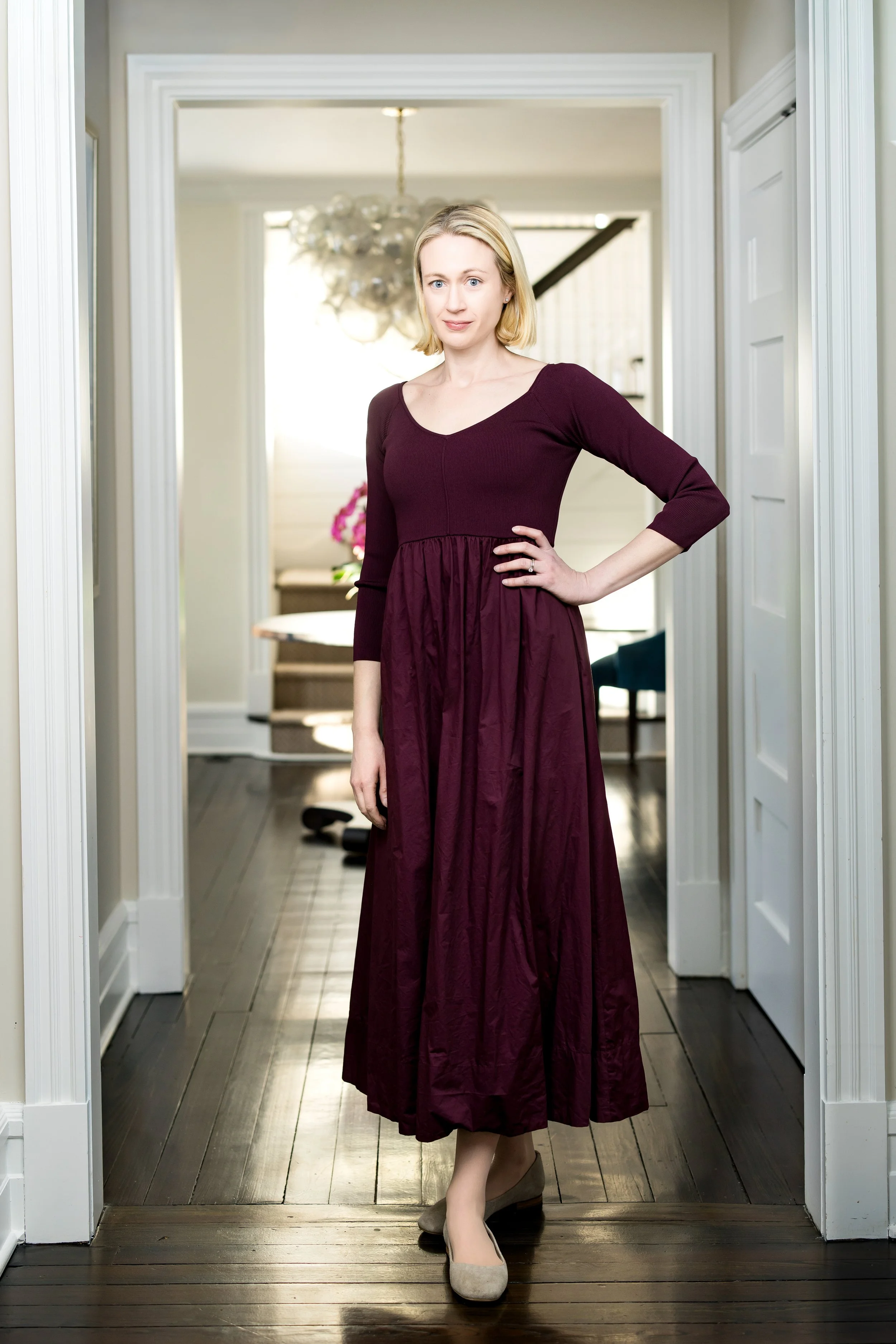 Full length picture of a woman with blonde hair in a burgundy dress standing in a doorway in her home