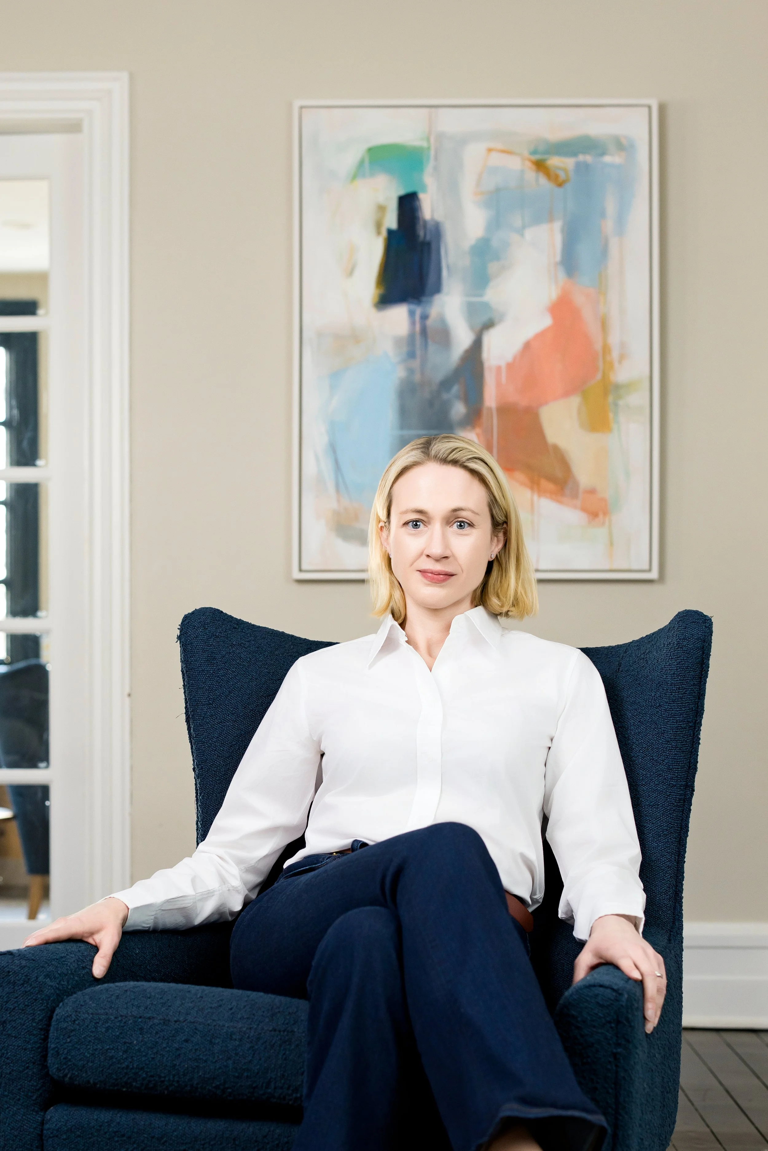 Woman with blonde hair and a white collar shirt leaned back in a blue chair with a painting behind her in her Westchester NY home