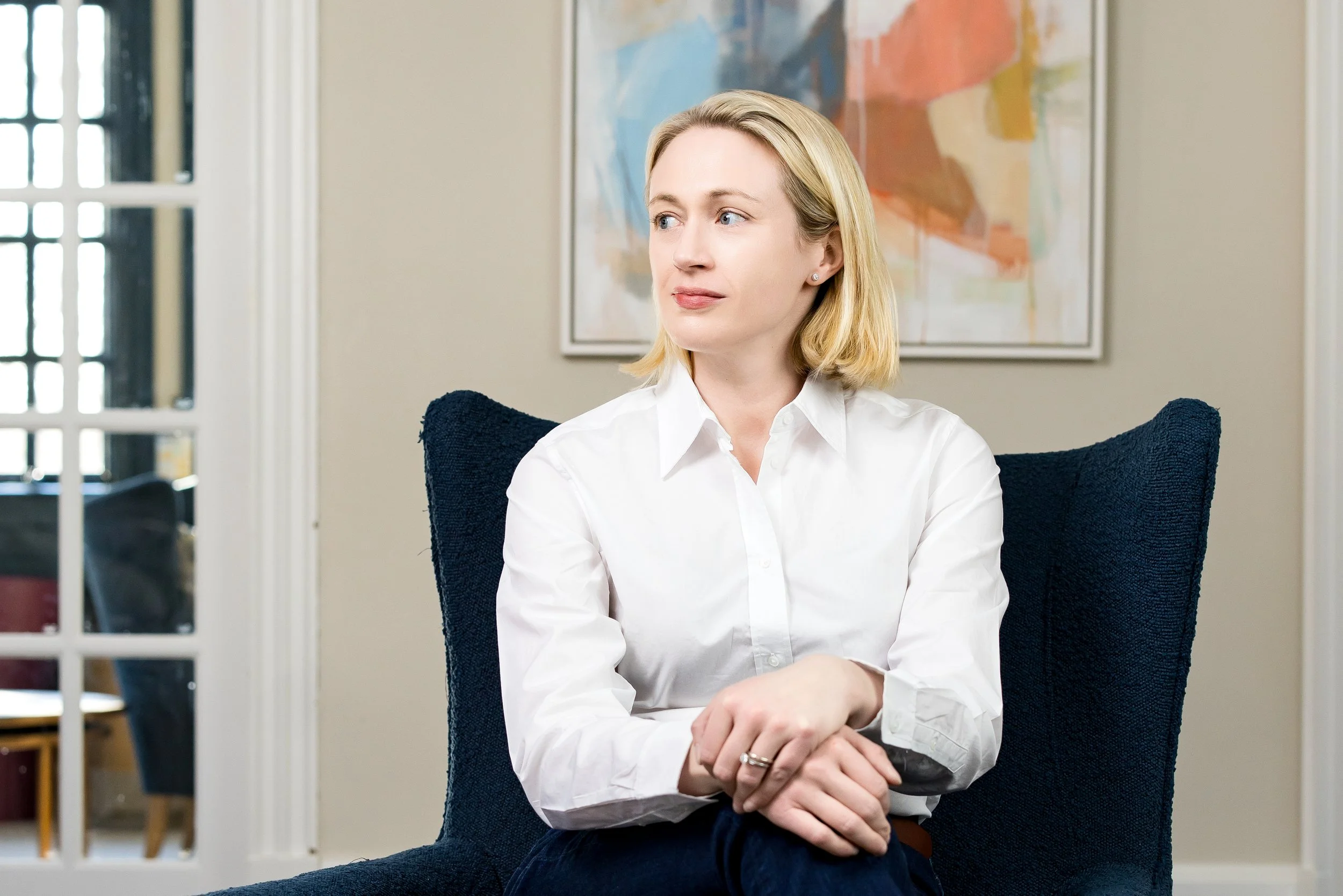woman with cropped blonde hair looking to the side seated in a chair in her home with a painting behind her