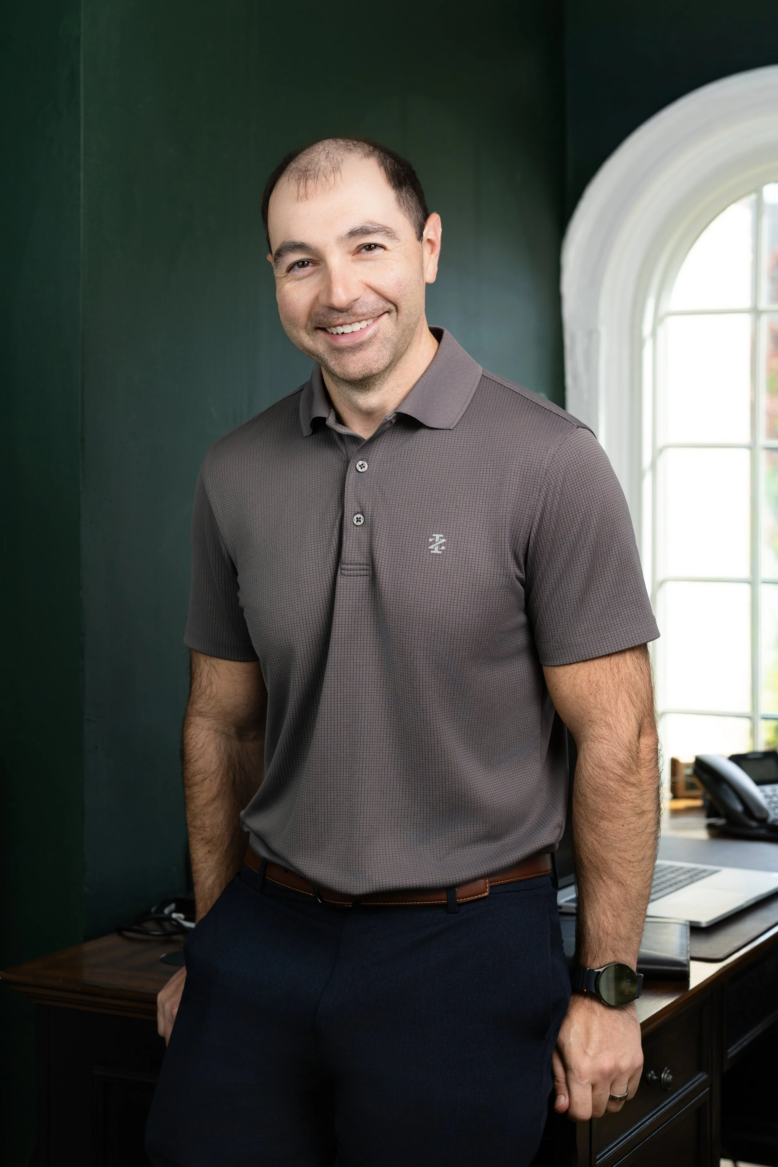 Headshot of a smiling man with a grey shirt standing in his office against his desk