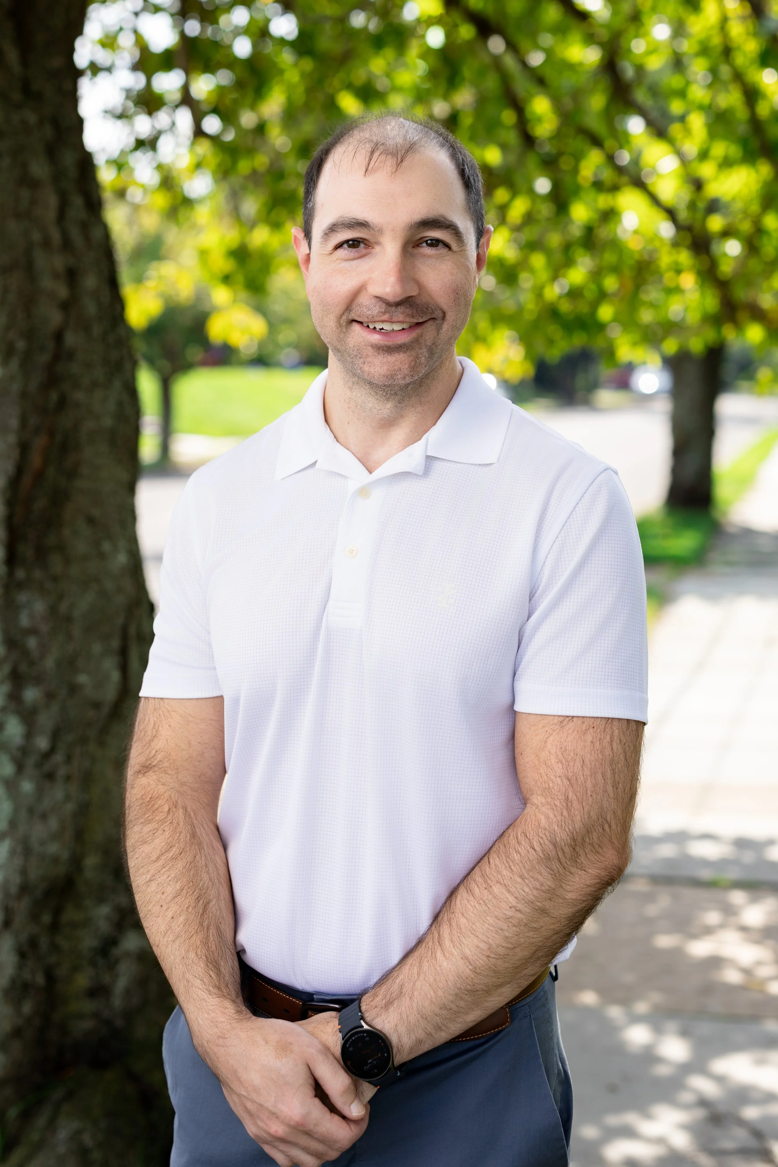 Outdoor headshot of a man in a white polo shirt in front of trees