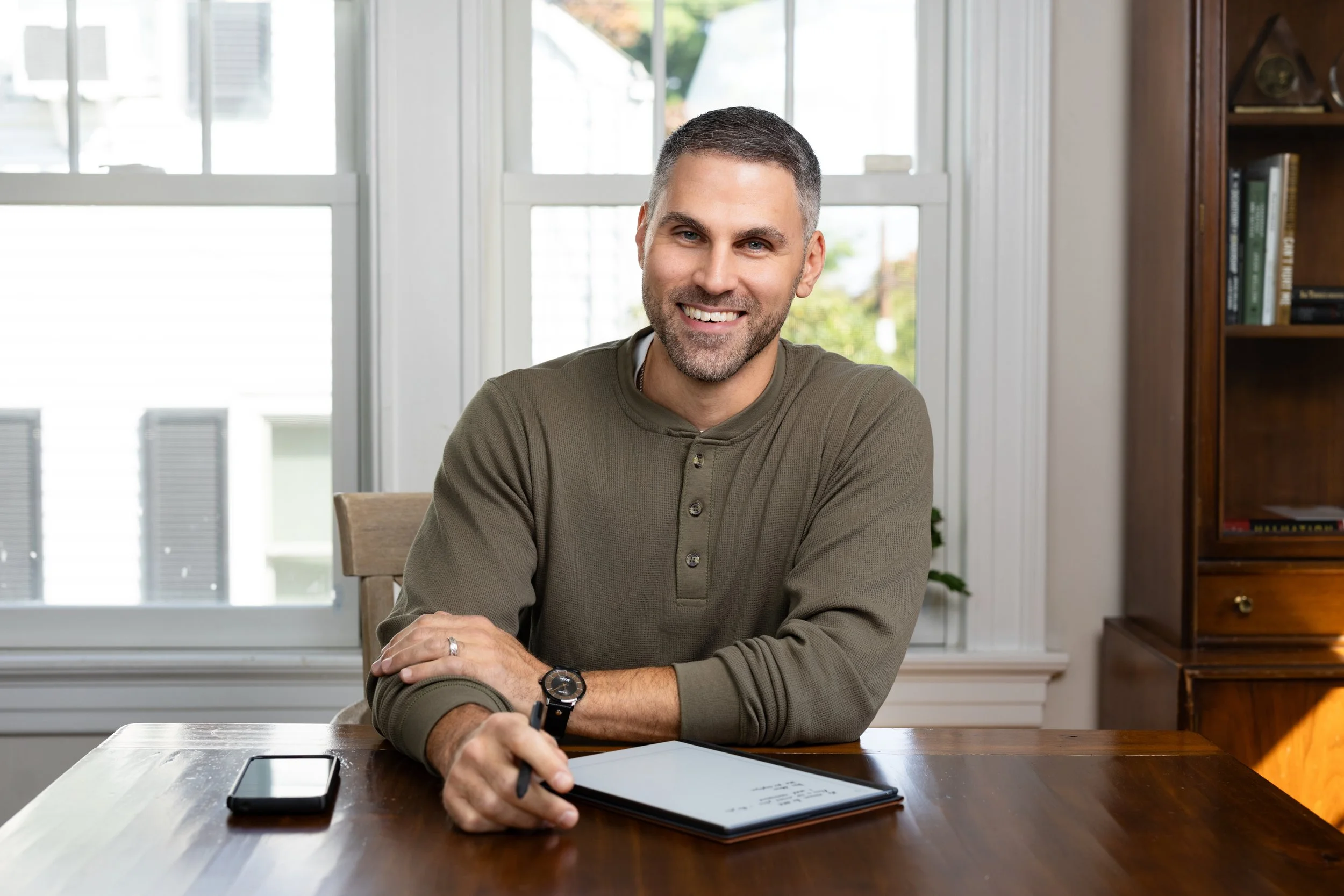 Man with grey hair and a brown shirt is seated at a desk smiling at the camera