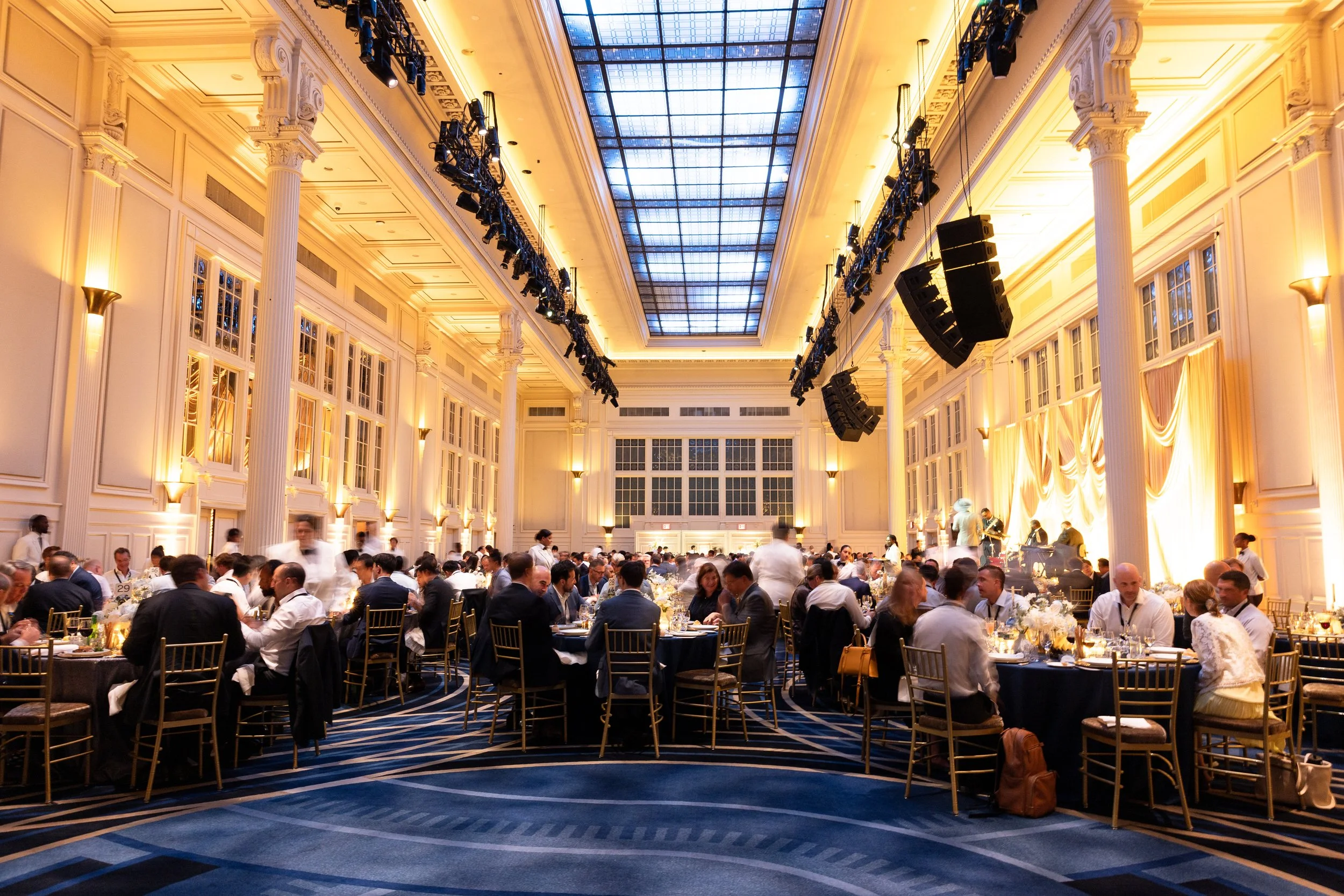 Ballroom setting with people seated at dinner in a room lit with yellow and a blue skylight