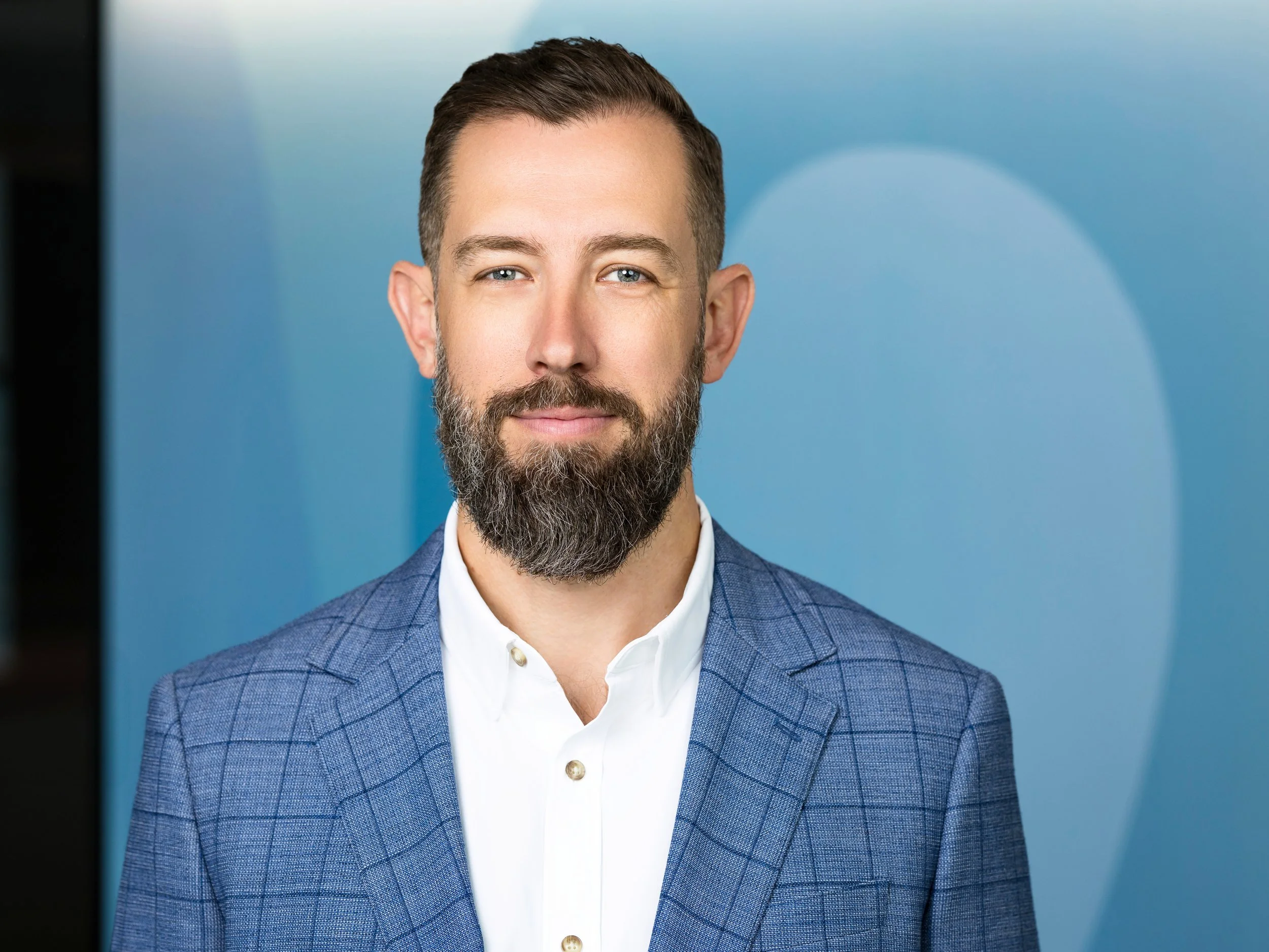 A headshot of a white man with a beard and blue jacket in an office setting