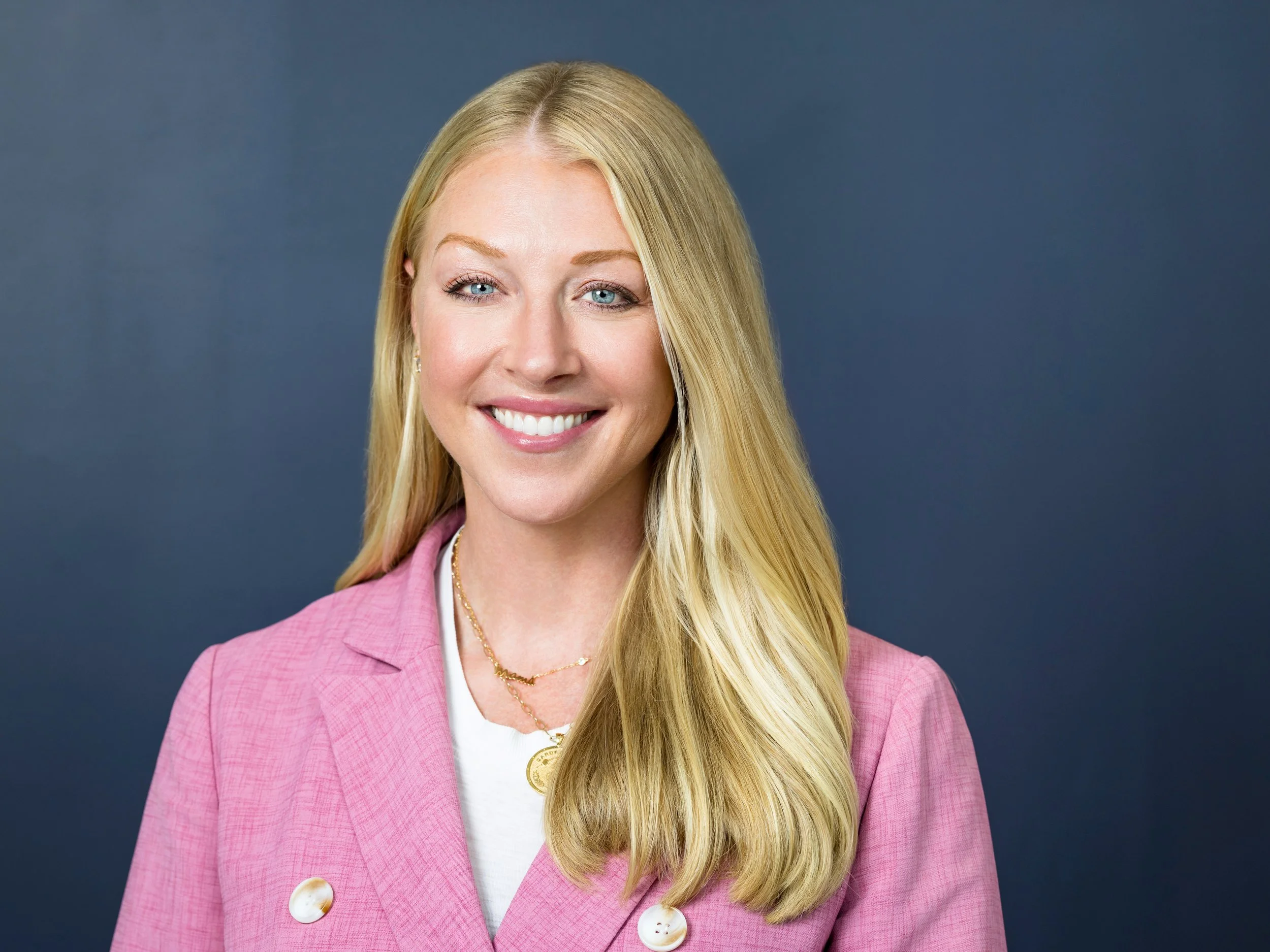 A headshot of smiling white woman with blonde hair and a pink jacket with a blue backdrop 