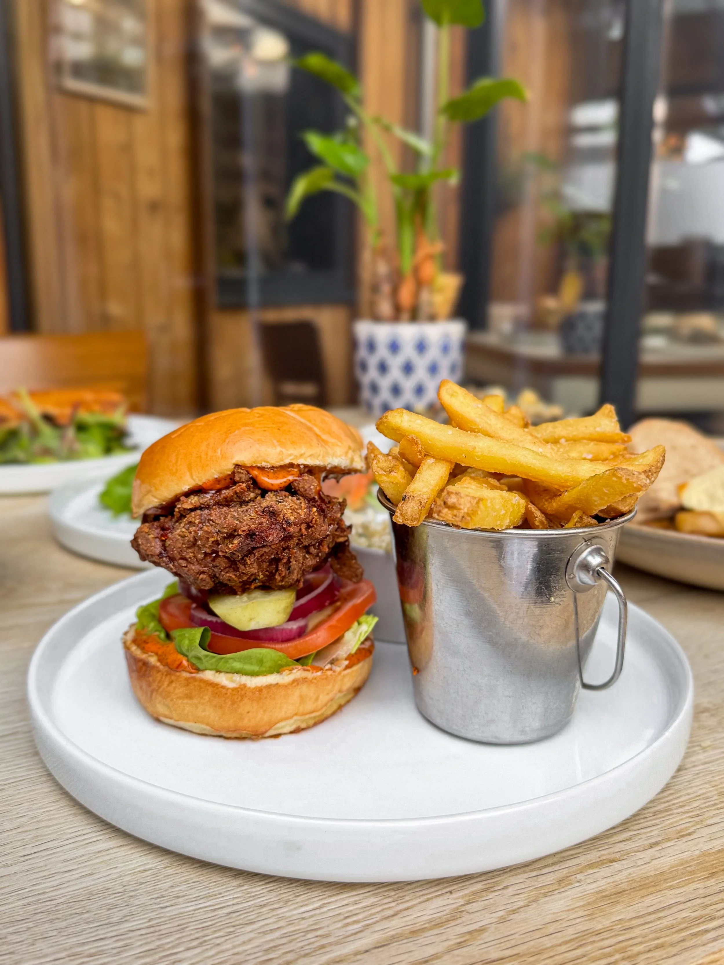 A plate with a burger containing lettuce, tomato, pickles, and a beef patty, accompanied by a metal cup of French fries, on a wooden table in a restaurant setting.