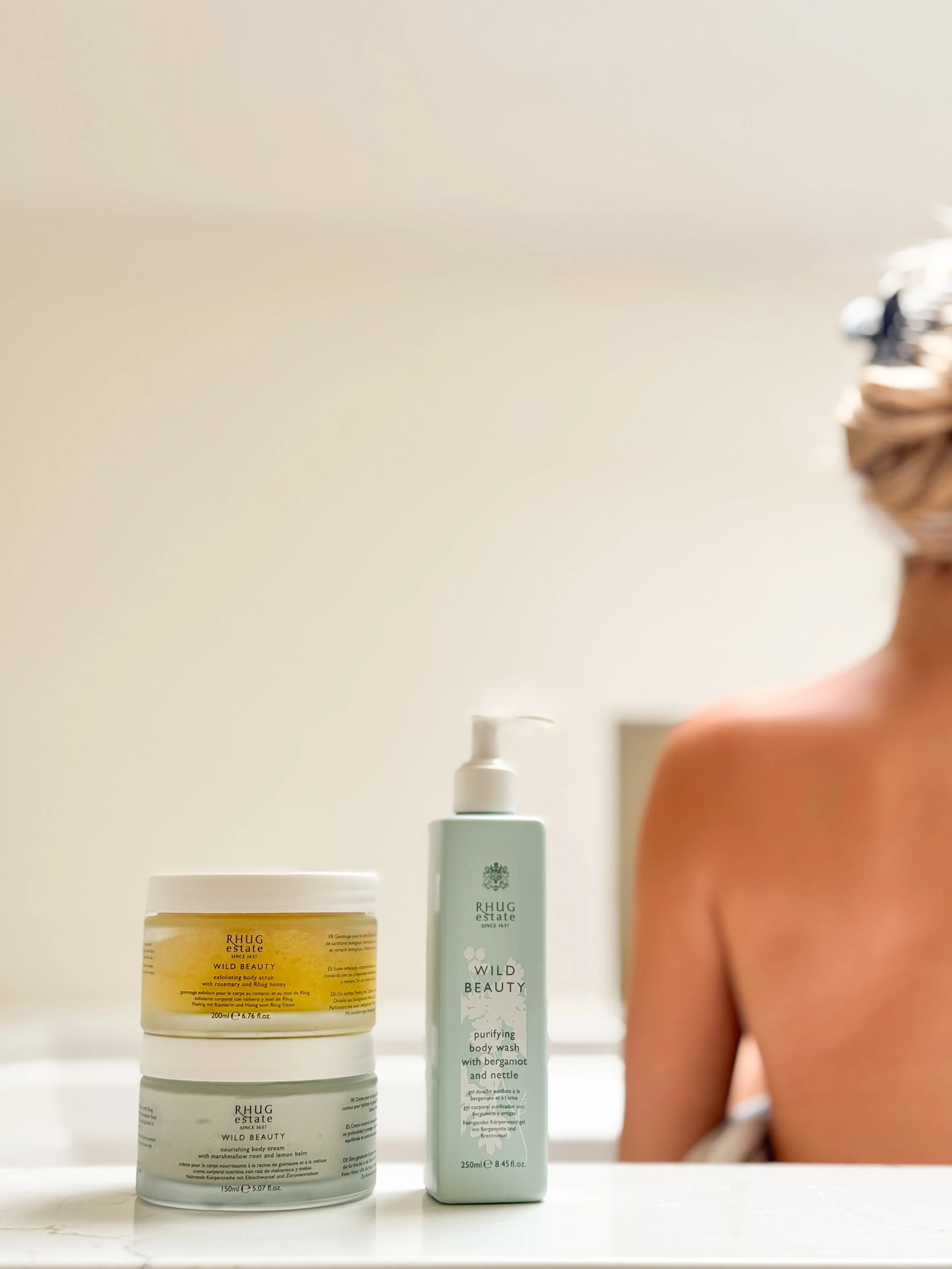 Collection of skincare and body care products from Rhug Estate, including a yellow jar labeled 'Wild Beauty' exfoliating body scrub, a white jar labeled 'Wild Beauty' nourishing body cream, and a white bottle of purifying body wash with bergamot and nettle, placed on a white surface with a blurred person in the background.