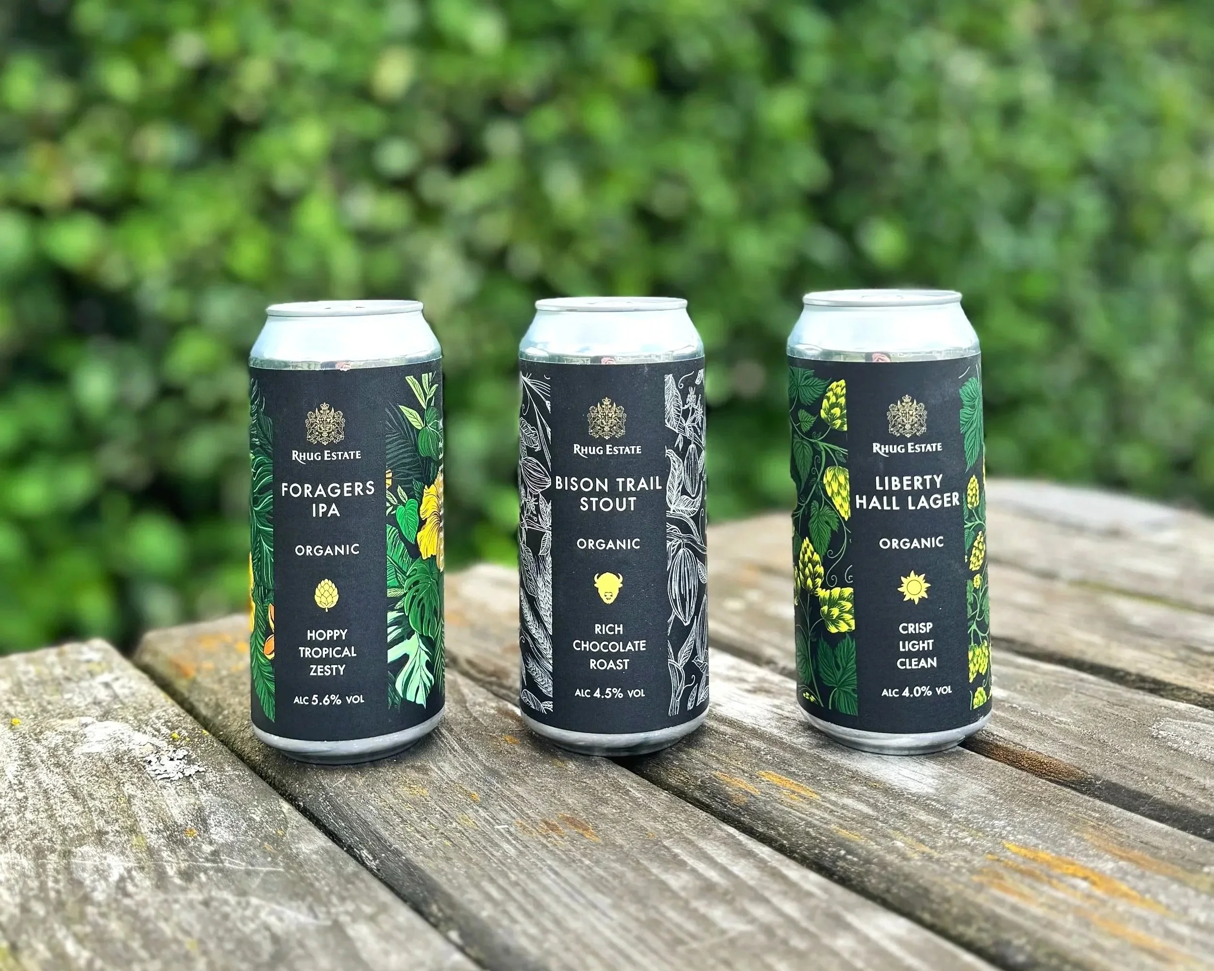 Three cans of craft beer from Rhug Estate, placed on a wooden surface outdoors. The cans feature black labels with lush green botanical illustrations and white text indicating the beer varieties: Foragers IPA, Bison Trail Stout, and Liberty Hall Lager, with alcohol contents of 5.6%, 4.5%, and 4.0% respectively.