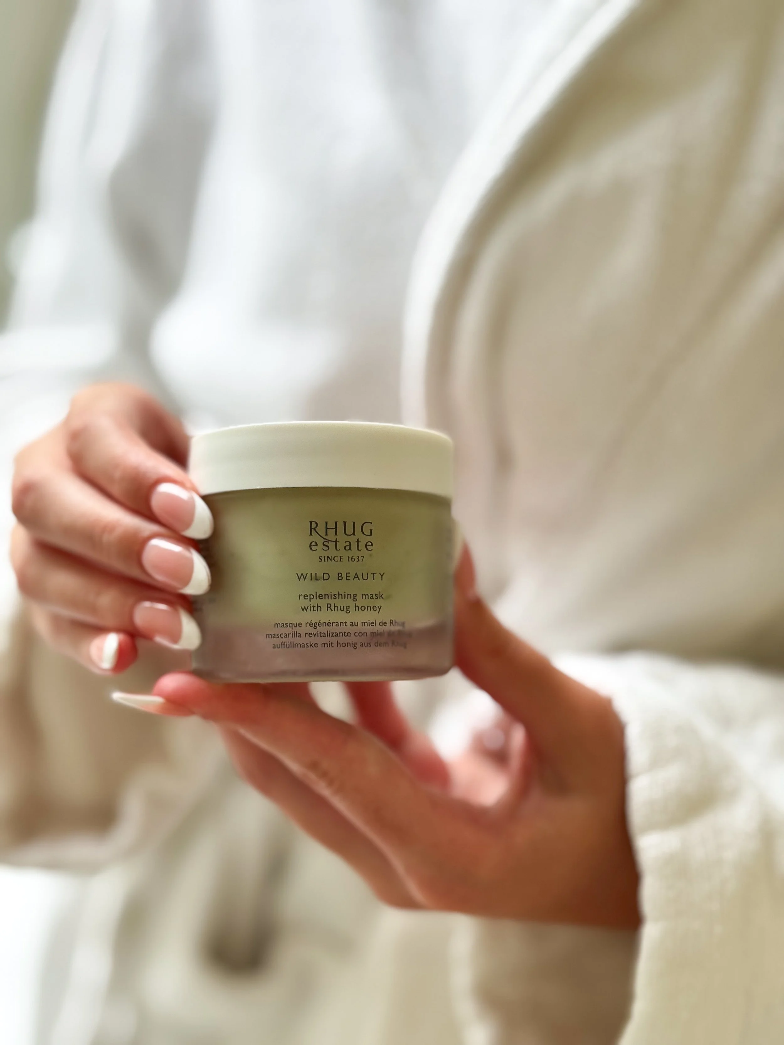 Person holding a beige container labeled 'RHUG estate' with additional text about a replenishing mask with Rhug honey, against a light-colored background.