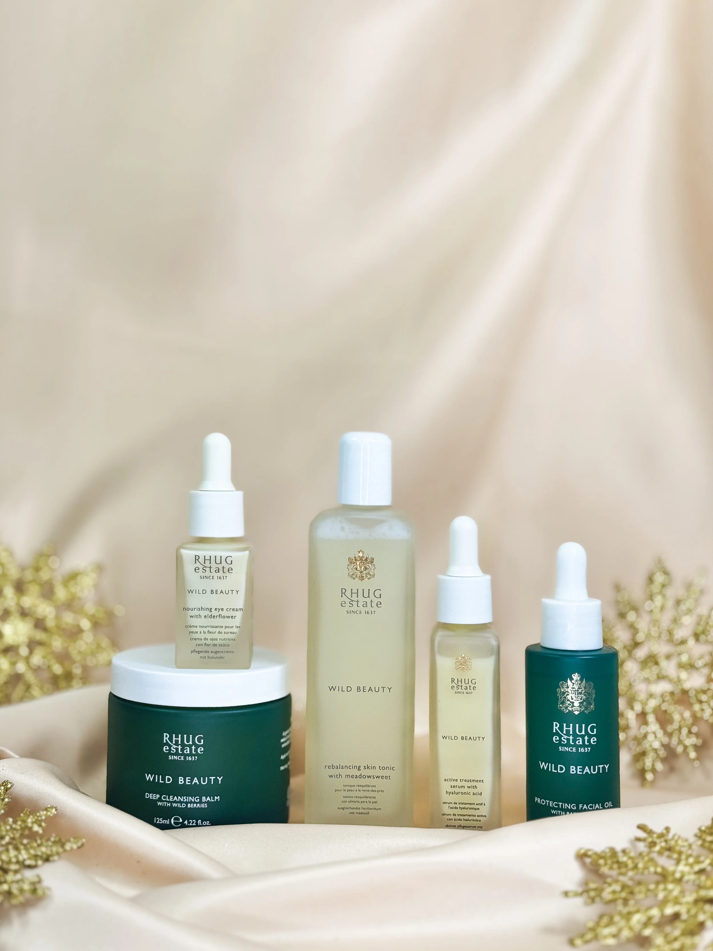 Collection of skincare products from Rhug Estate, including cleansing balm, toning liquid, serum, and facial oil, arranged on a satin fabric with gold decorative elements.