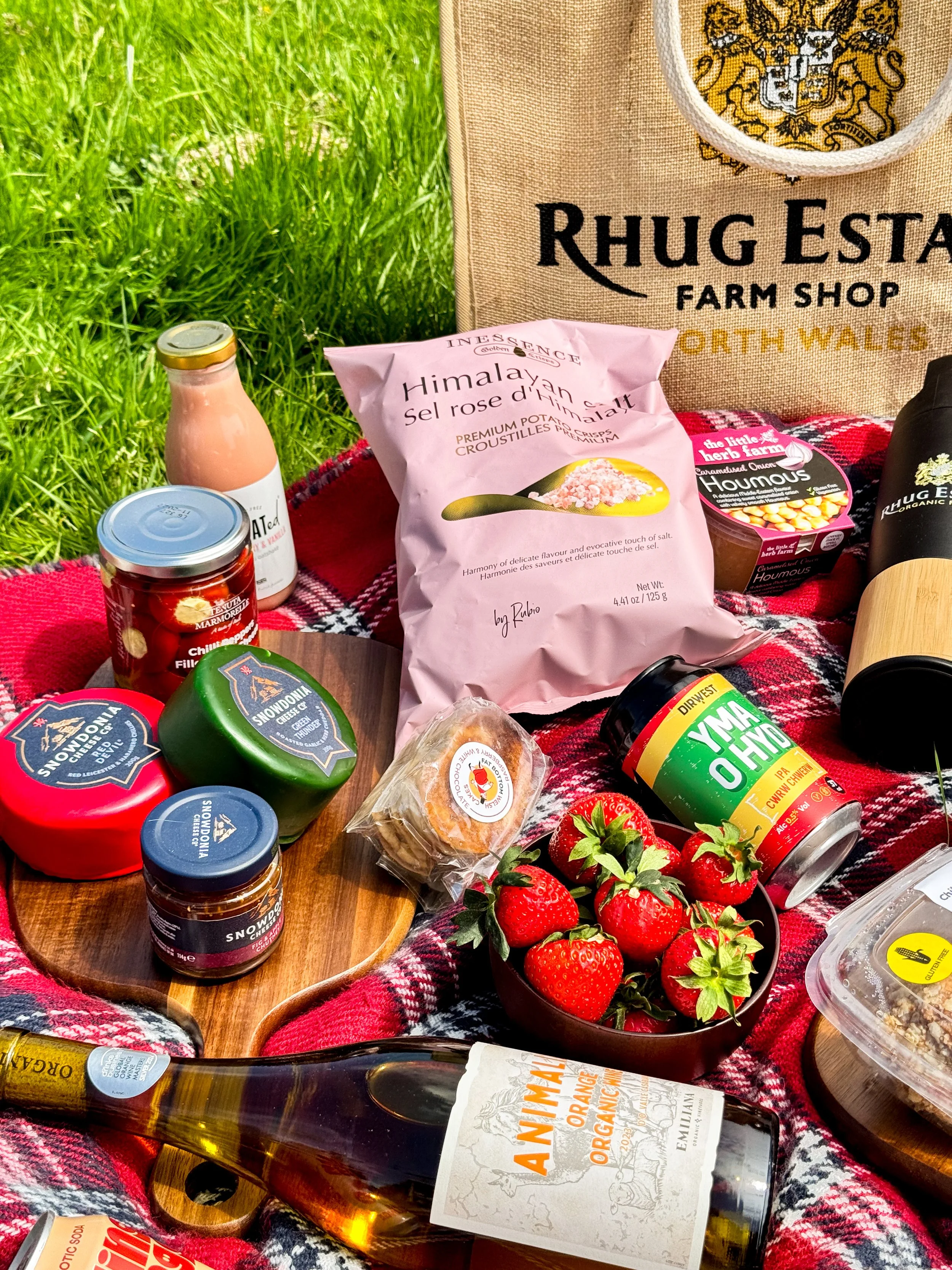 A picnic scene with various food items on a red plaid blanket on the grass, including strawberries, canned goods, a bottle of organic orange juice, a bag of Himalayan salt, and a bag from Rhug Estate Farm Shop, with a wooden tray and a magazine.