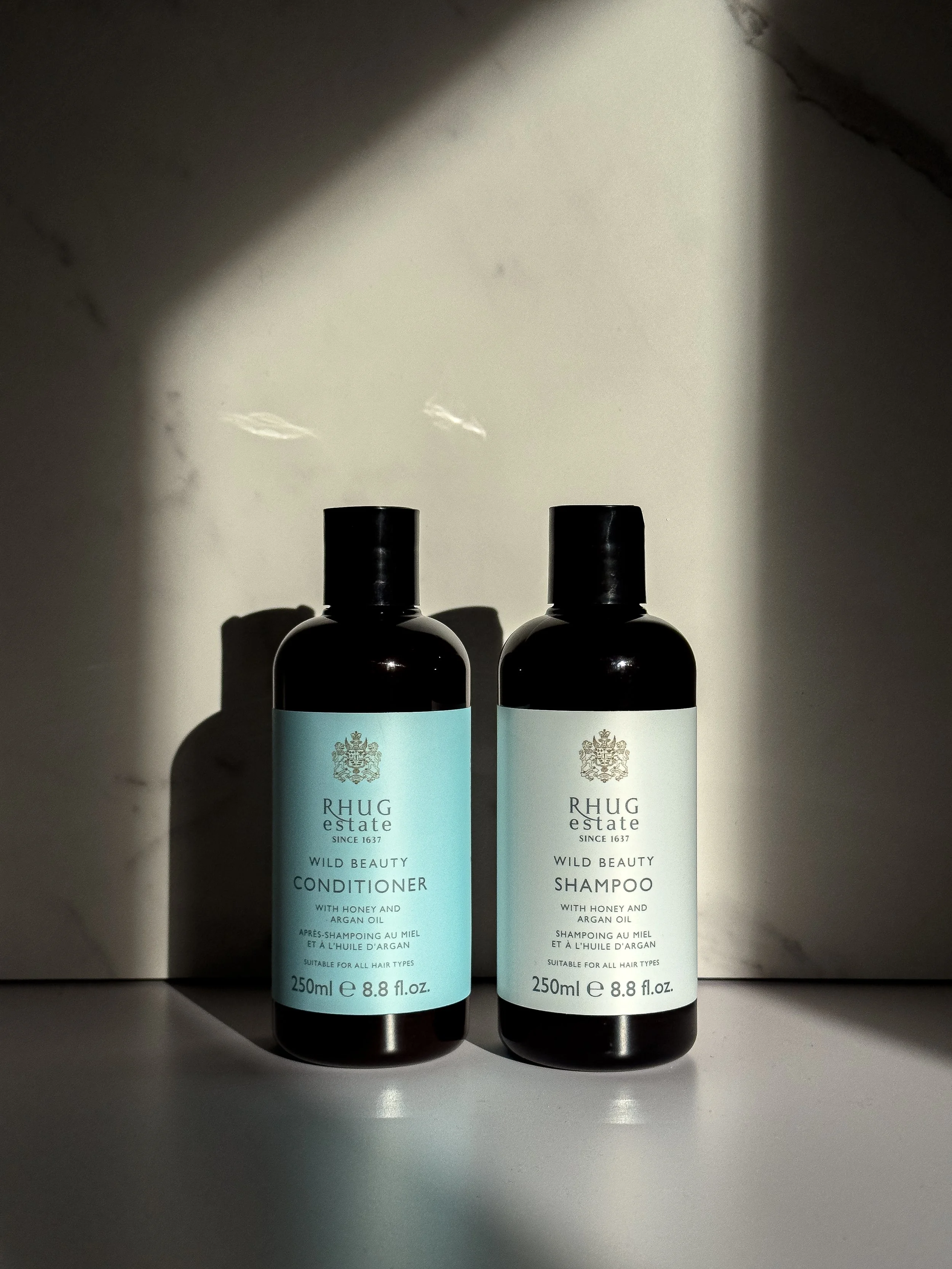 Two bottles of RHUG estate Wild Beauty hair care products, one labeled conditioner and the other shampoo, stand side by side on a surface with shadows partially covering them.