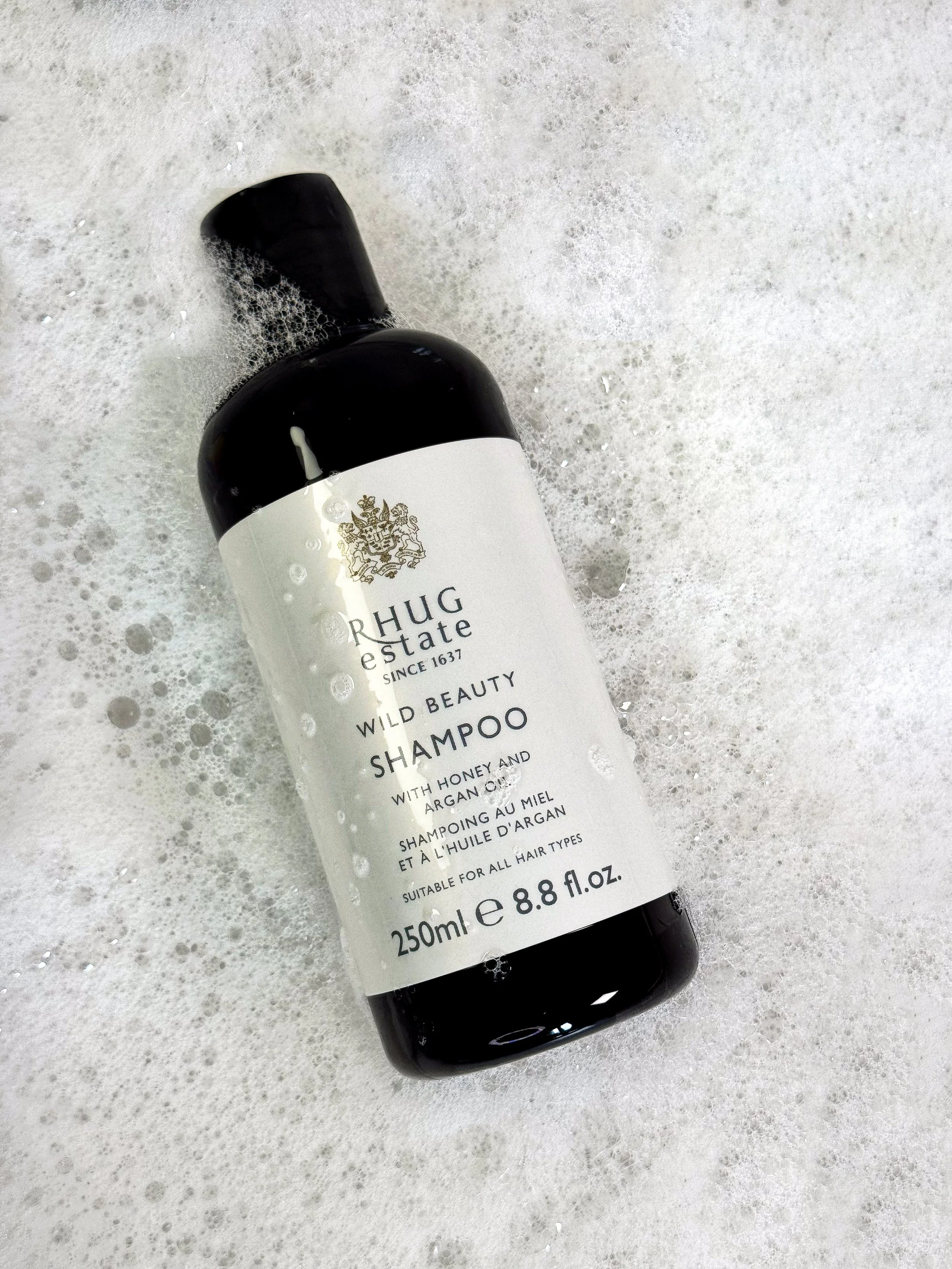 A black bottle of shampoo with a white label, lying on a foamy, bubbly surface.