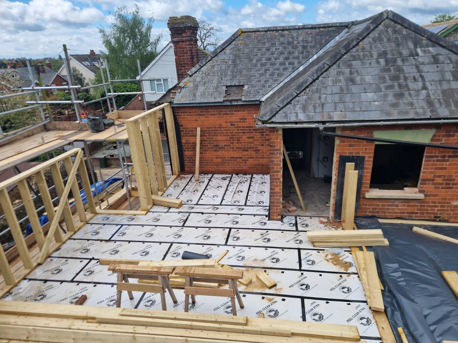 Construction site on a rooftop with wooden framing and a partially built balcony, with brick houses and trees in the background.
