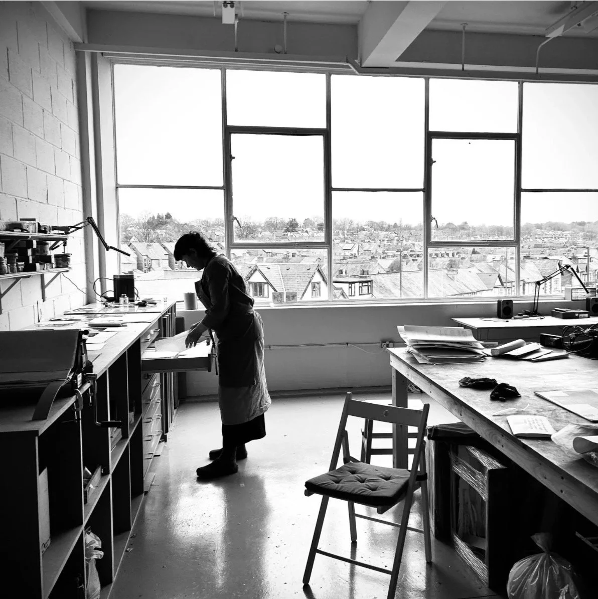 A woman in an art studio or workshop working at a desk near large windows overlooking a cityscape.