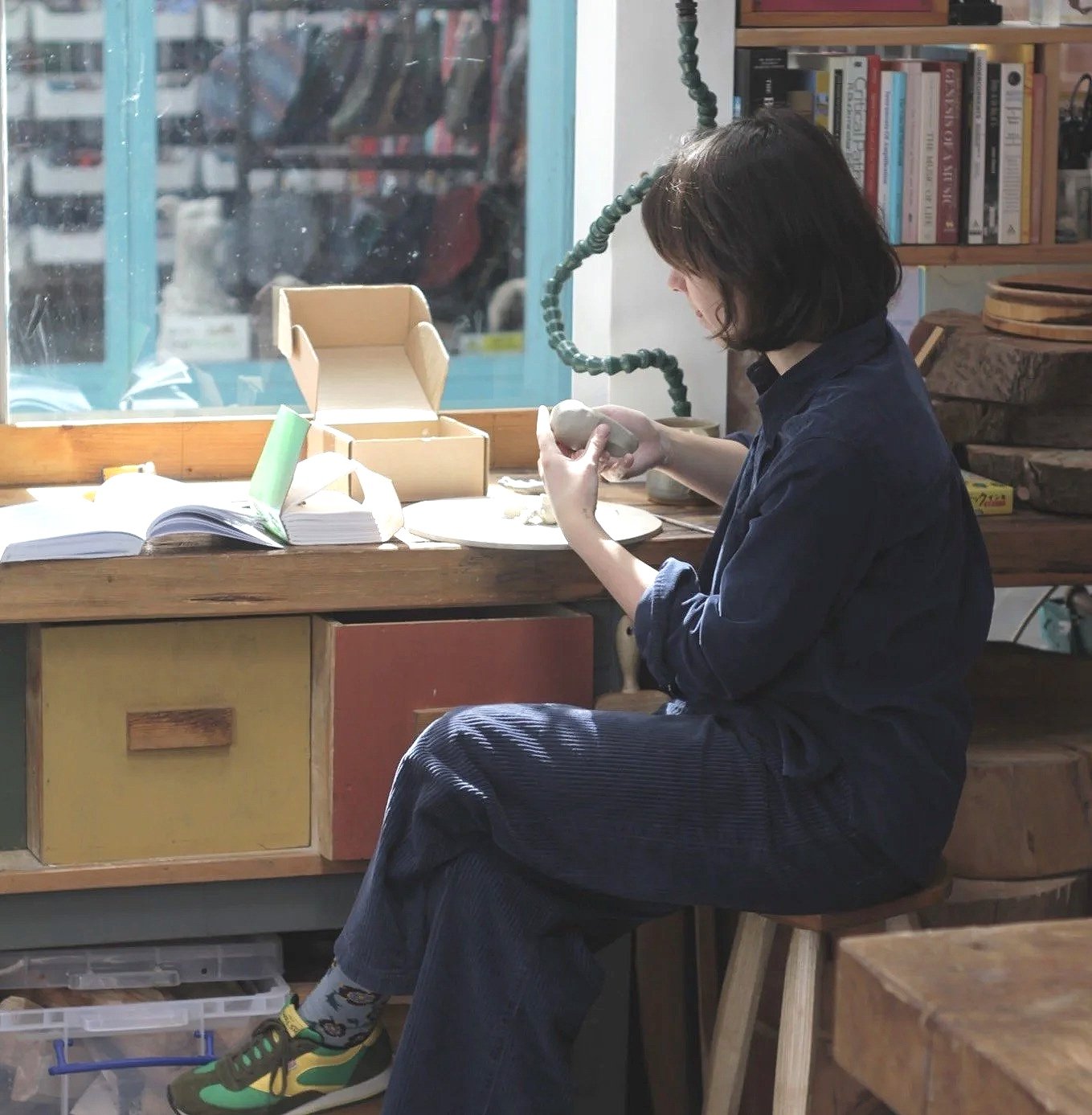 A woman with shoulder-length dark hair sitting on a wooden stool, working on a small sculpture or clay object at a wooden work table in a cozy room filled with bookshelves, a window, and various art supplies.