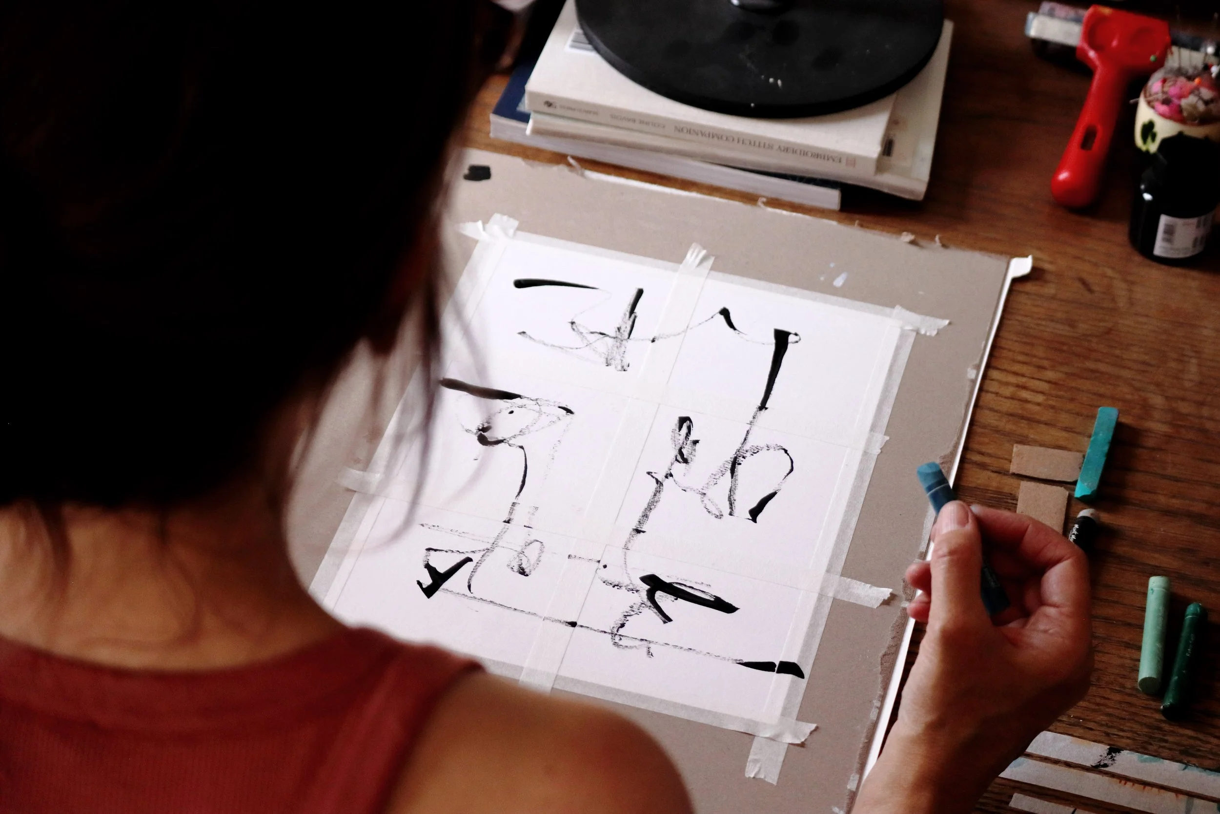 Person creating abstract black ink drawings on paper with colored markers, sitting at a wooden table with art supplies.