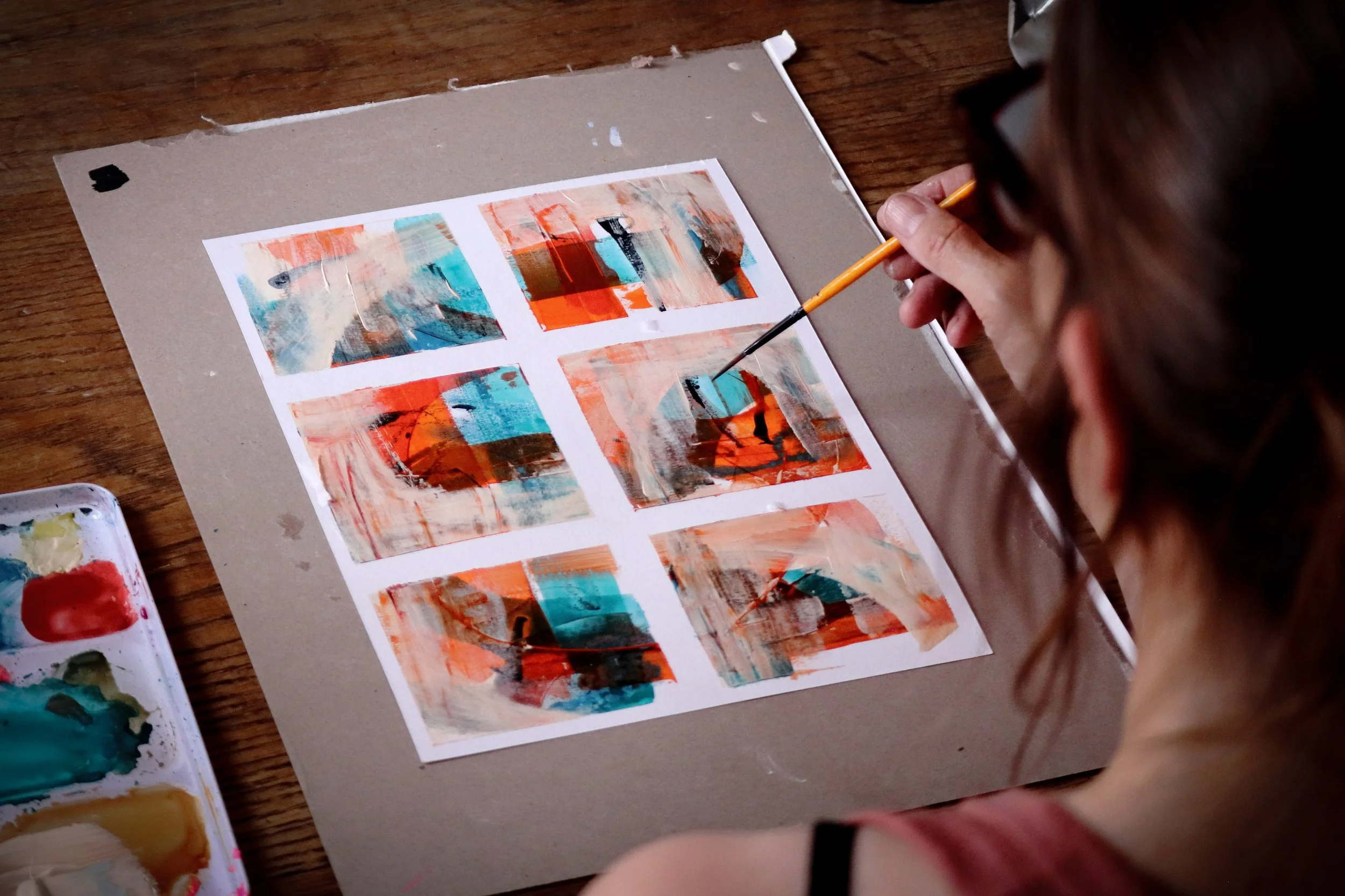 Person creating abstract paintings with orange, blue, black, and beige brushstrokes on a sheet of paper, placed on a wooden table, with watercolor paints nearby.